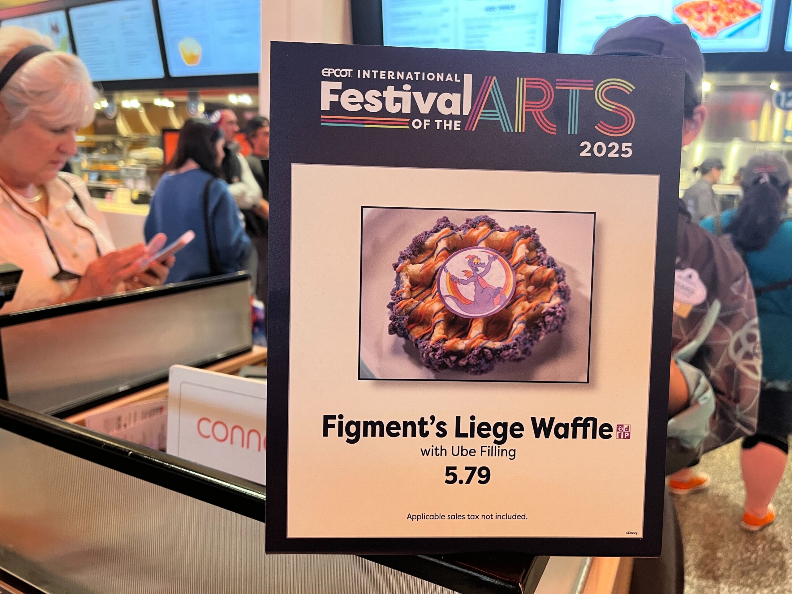 A sign for "Figment's Liege Waffle with Ube Filling" priced at $5.79 stands prominently at the Epcot International Festival of the Arts 2025. In the backdrop, vibrant menu boards and lively festival-goers animate the bustling atmosphere.