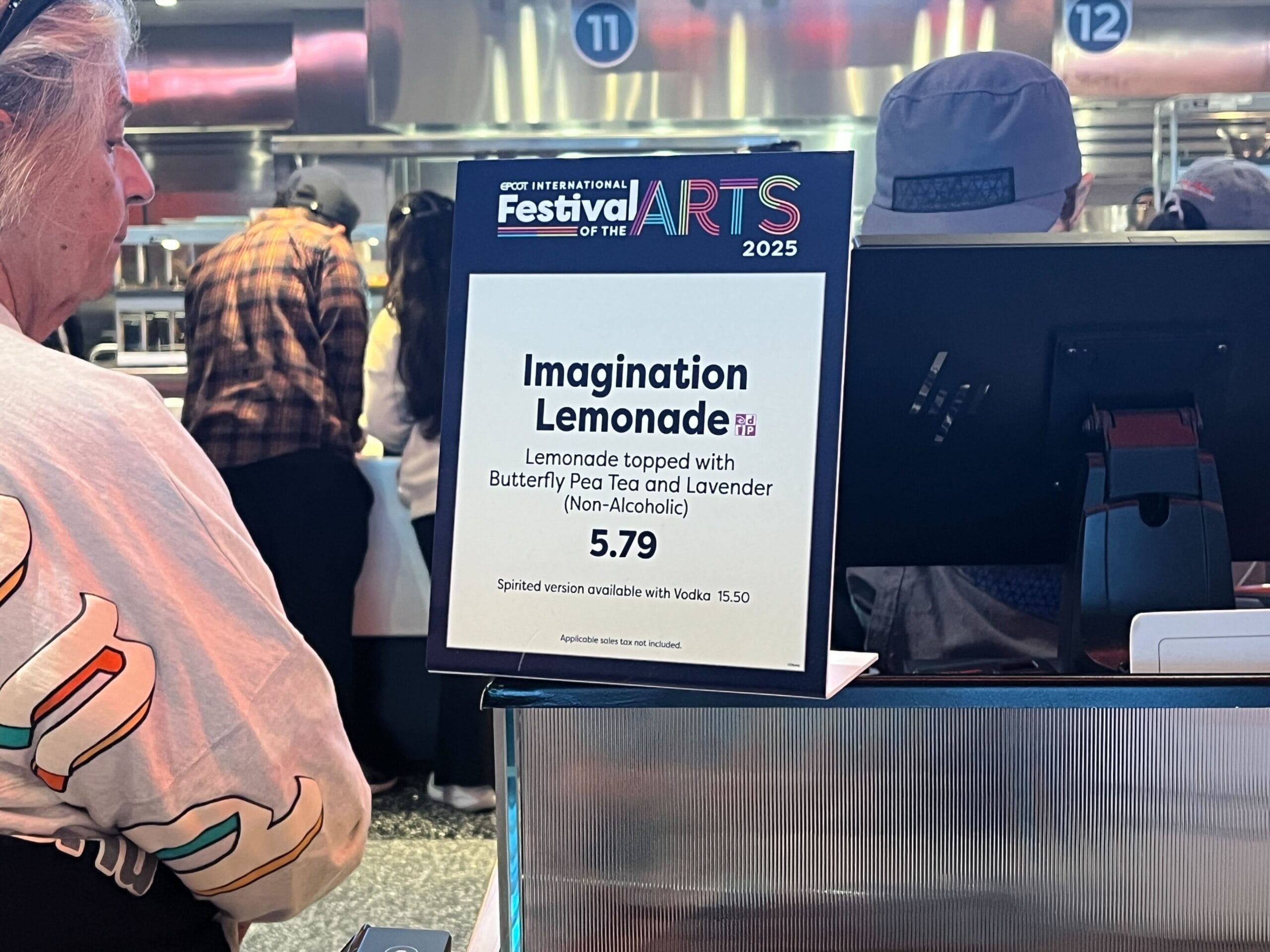 At the Festival of the Arts, a sign displays "Imagination Lemonade" with butterfly pea tea and lavender for $5.79. There's an option to add vodka for $15.50, while a person stands nearby, soaking in the creative atmosphere.