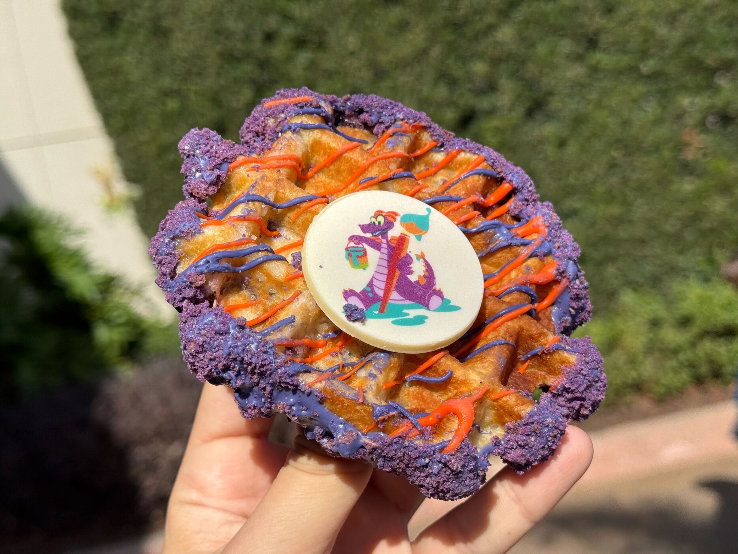 A vibrant pastry adorned with purple and orange icing, crowned by a circular white candy displaying a cartoon dragon, is cradled in someone's hand against a lush backdrop of foliage—perfect for celebrating the Festival of the Arts.