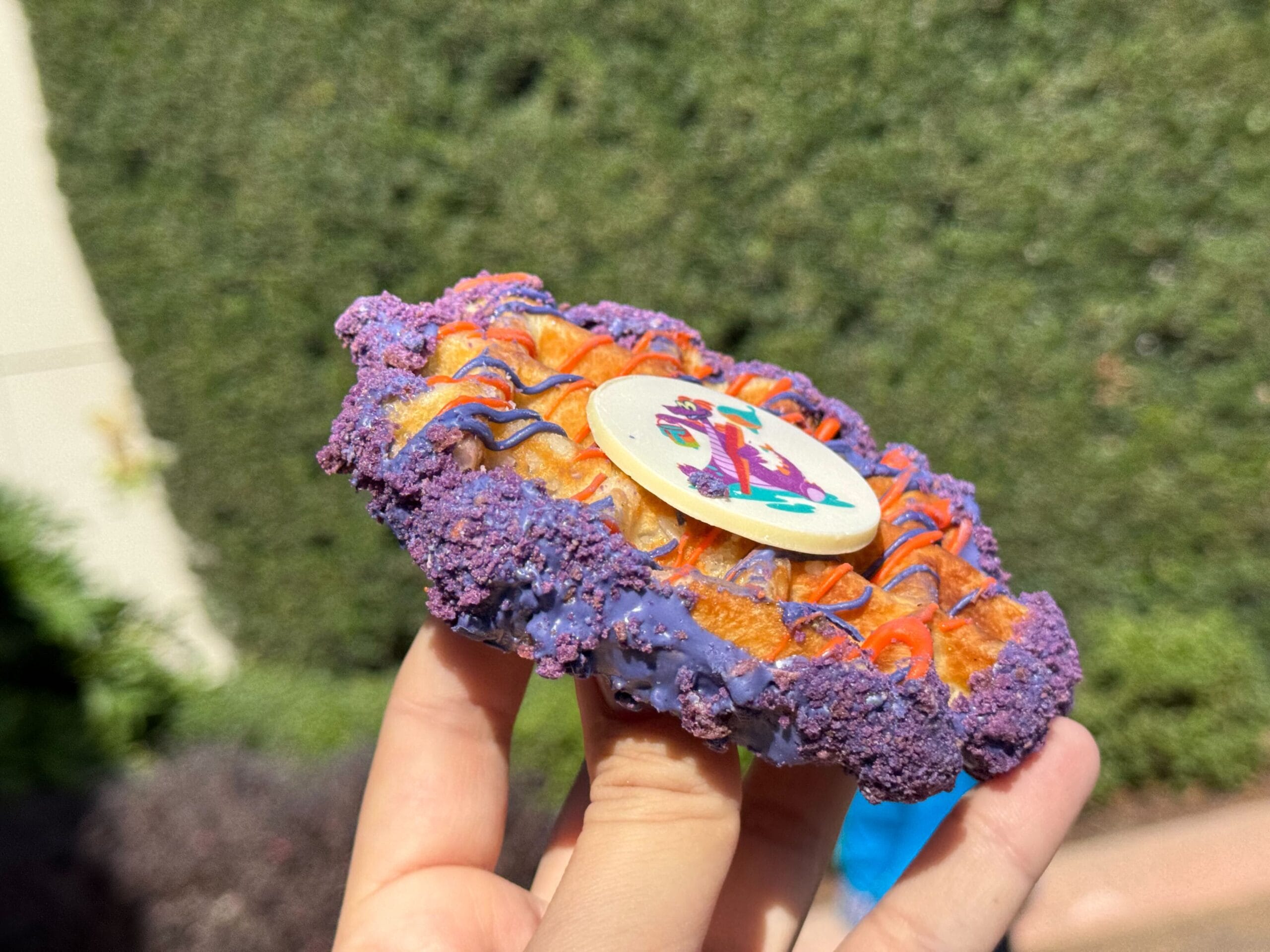 Close-up of a hand holding a colorful, round pastry with a decorated top, reminiscent of the vibrant creations at the Festival of the Arts, featuring purple crumbles and orange icing. The background showcases an out-of-focus hedge.