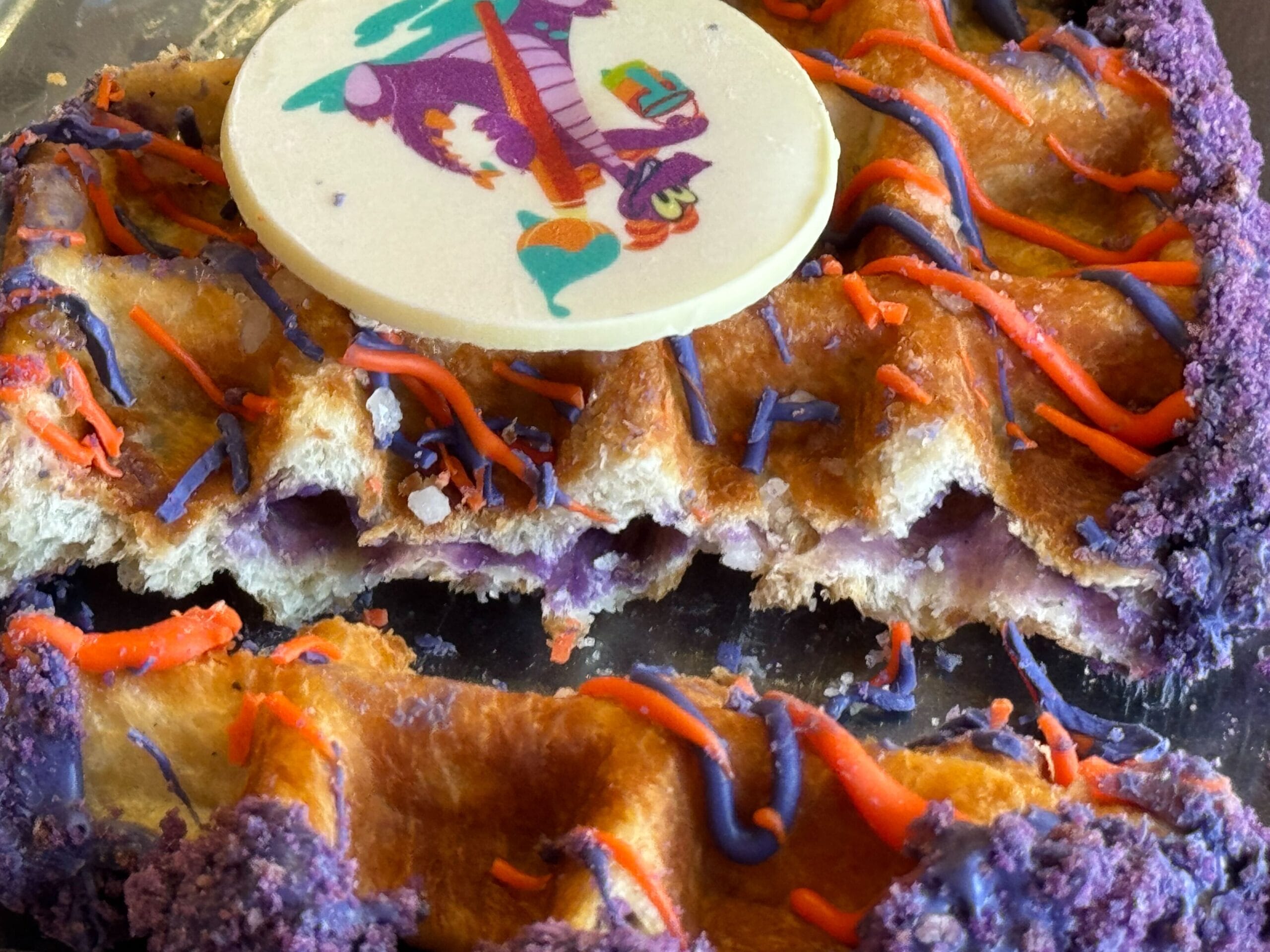 A waffle adorned with purple and orange icing, a round edible unicorn decoration, and purple sprinkles gracefully edging the treat, reminiscent of the vibrant creativity at the Festival of the Arts.