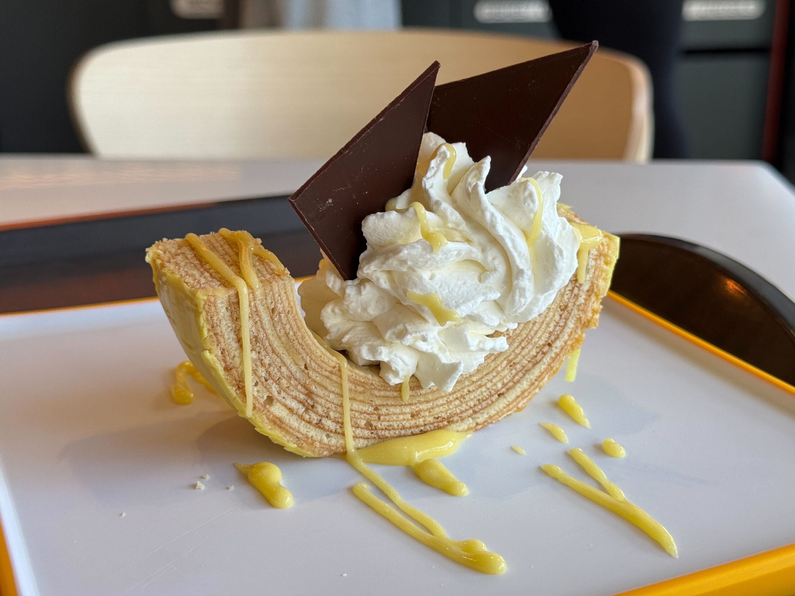 At the Festival of the Arts, indulge in a pastry slice adorned with whipped cream and twin chocolate triangles, elegantly drizzled with a zesty yellow sauce, all served on a pristine white plate accented by an orange rim.