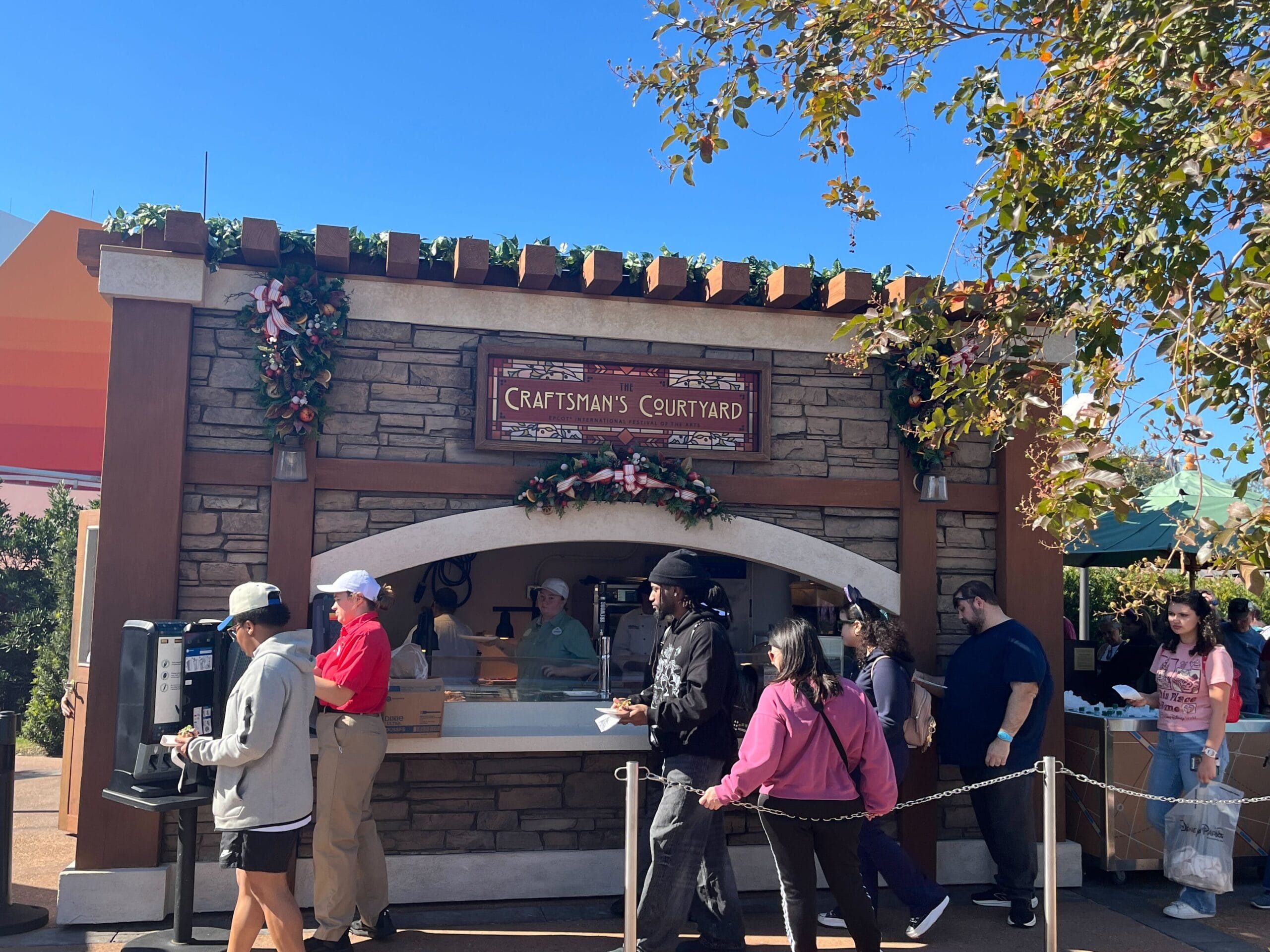 People standing in line at the Festival of the Arts food booth, labeled "Craftsman’s Courtyard," adorned with greenery and pink ribbons.