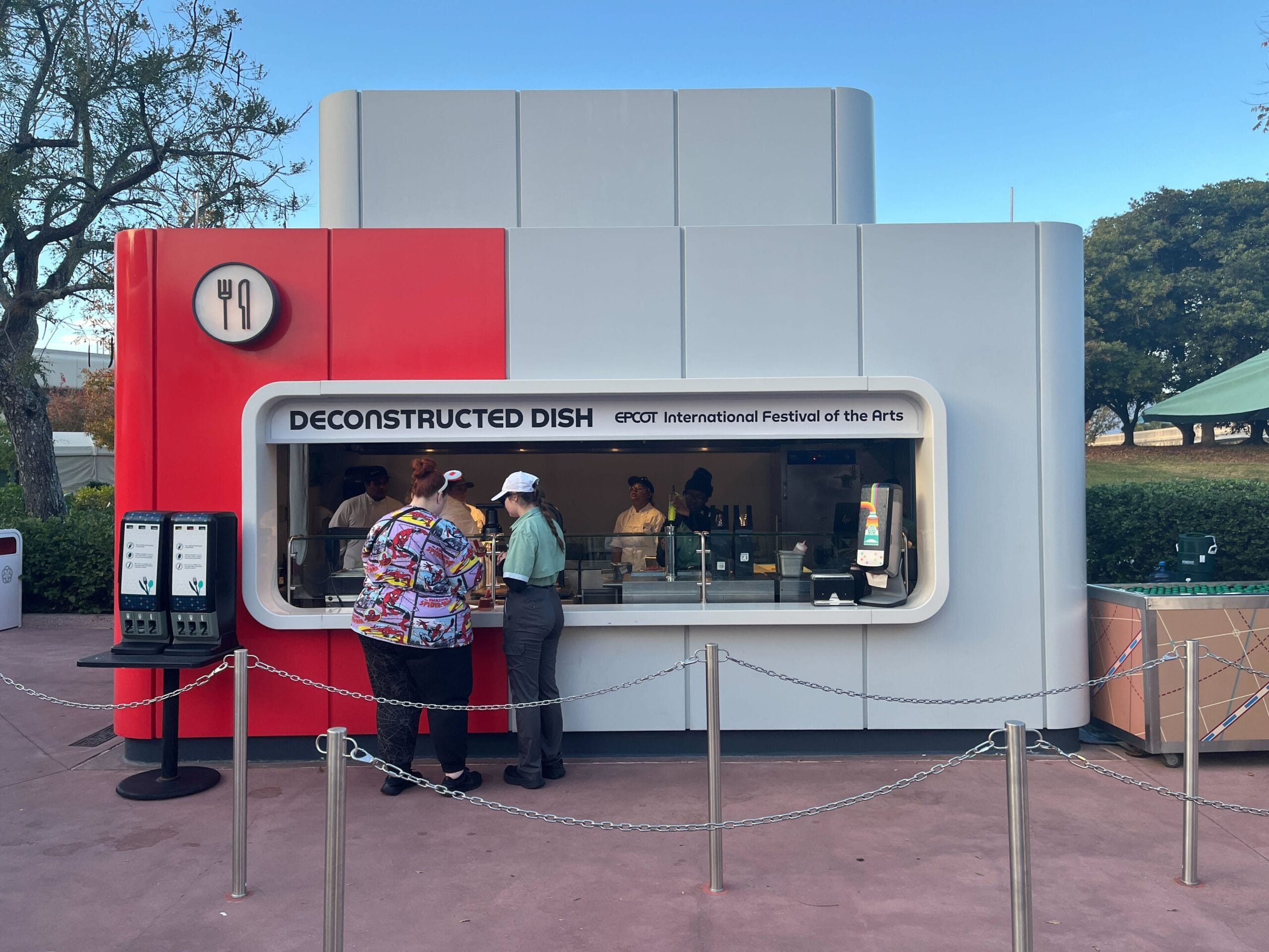 A small kiosk labeled "DECONSTRUCTED DISH" at the vibrant EPCOT International Festival of the Arts, bustling with people eagerly ordering at the counter.