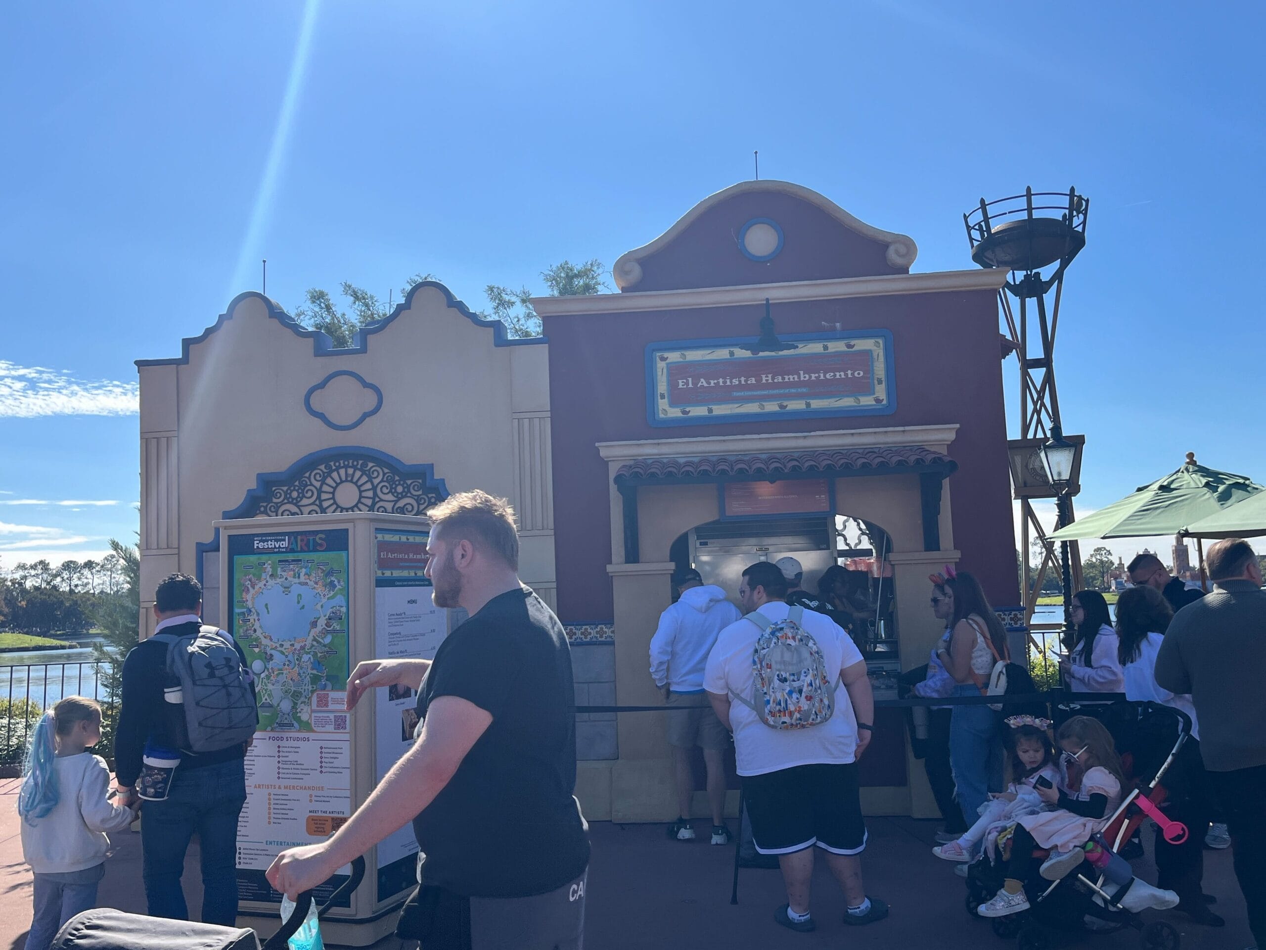 People stand in line at a food kiosk labeled "El Artista Hambriento" under the sun's warm embrace during the bustling Festival of the Arts. A man with a stroller and map surveys the scene in the foreground, planning his next artistic adventure.