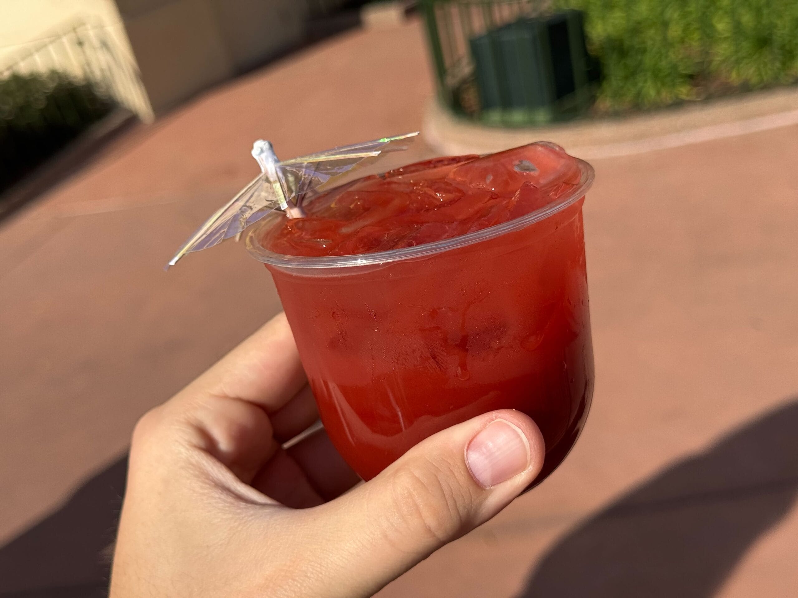 A hand holds a plastic cup with a red drink and ice, topped with a small decorative umbrella, against an outdoor background at the Festival of the Arts.