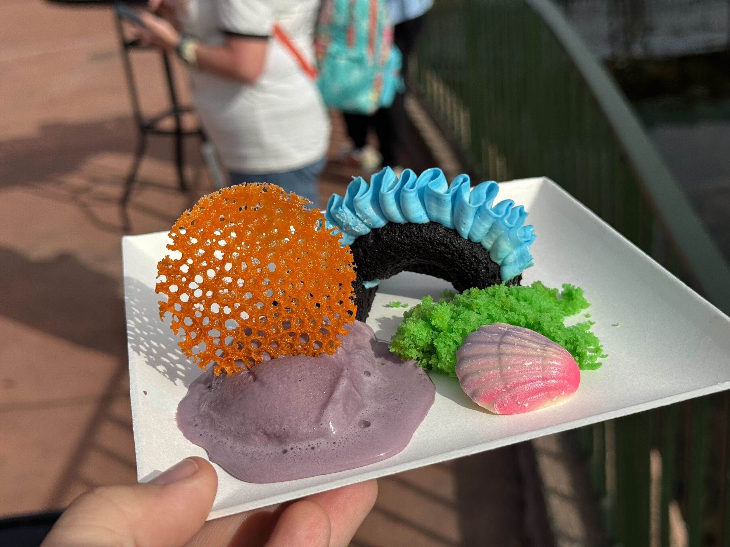 A plate of colorful, artistic desserts, reminiscent of the Festival of the Arts, features an orange tuile, a pink seashell-shaped delight, blue frosting, and green crumbles, all set against a backdrop of people enjoying the vibrant atmosphere.