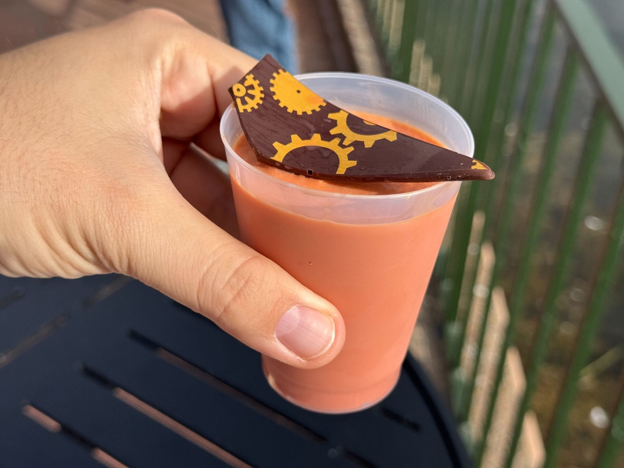 A person holds a plastic cup filled with pink-orange liquid, topped with a piece of chocolate decorated with yellow gear-shaped patterns, reminiscent of the creative flair found at the Festival of the Arts.