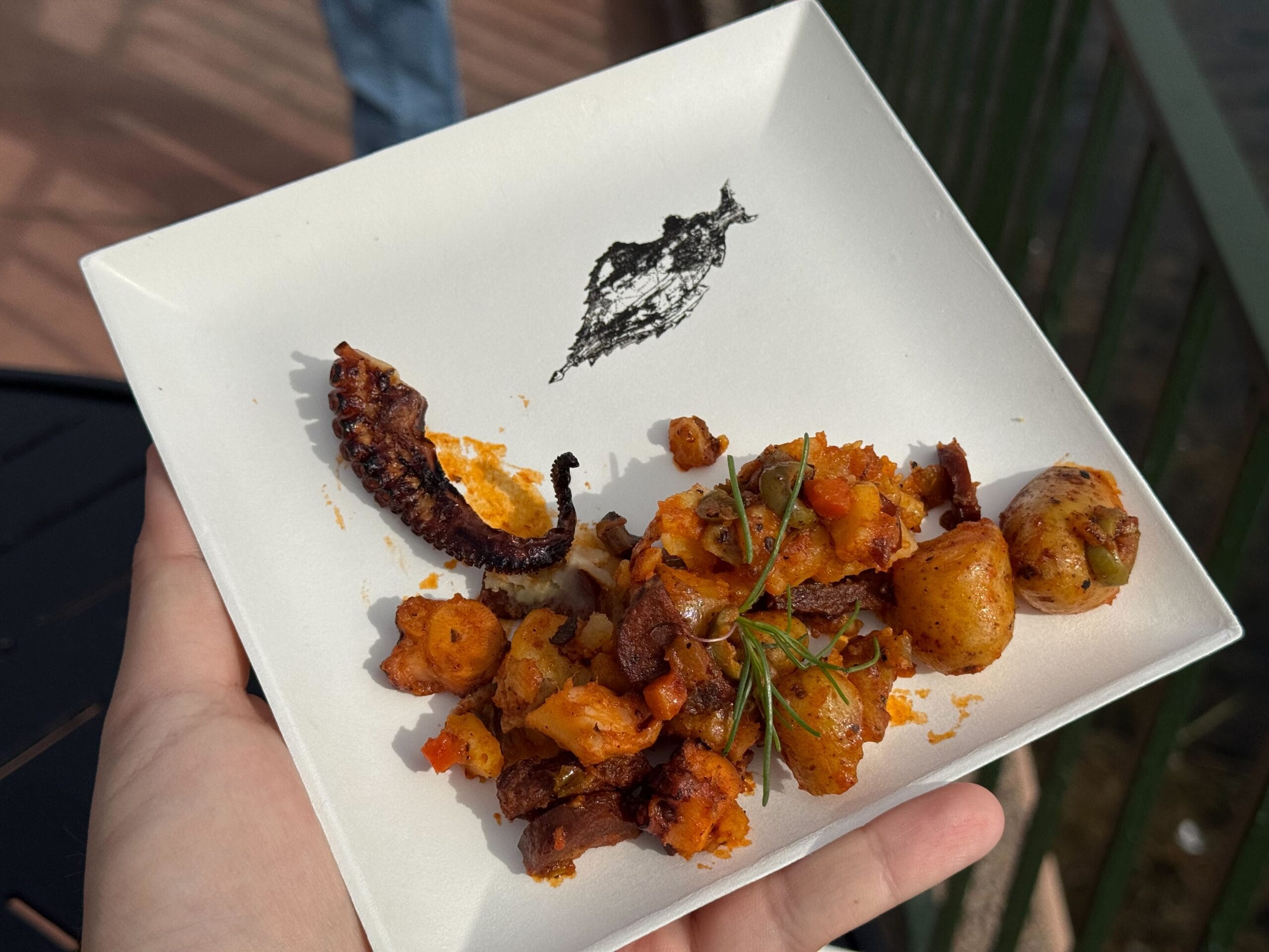 A plate featuring a perfectly grilled octopus tentacle, assorted roasted vegetables, and fresh herbs is showcased in someone's hand. Against a backdrop of outdoor railing and floor, this culinary masterpiece is a true testament to the creativity celebrated at the Festival of the Arts.