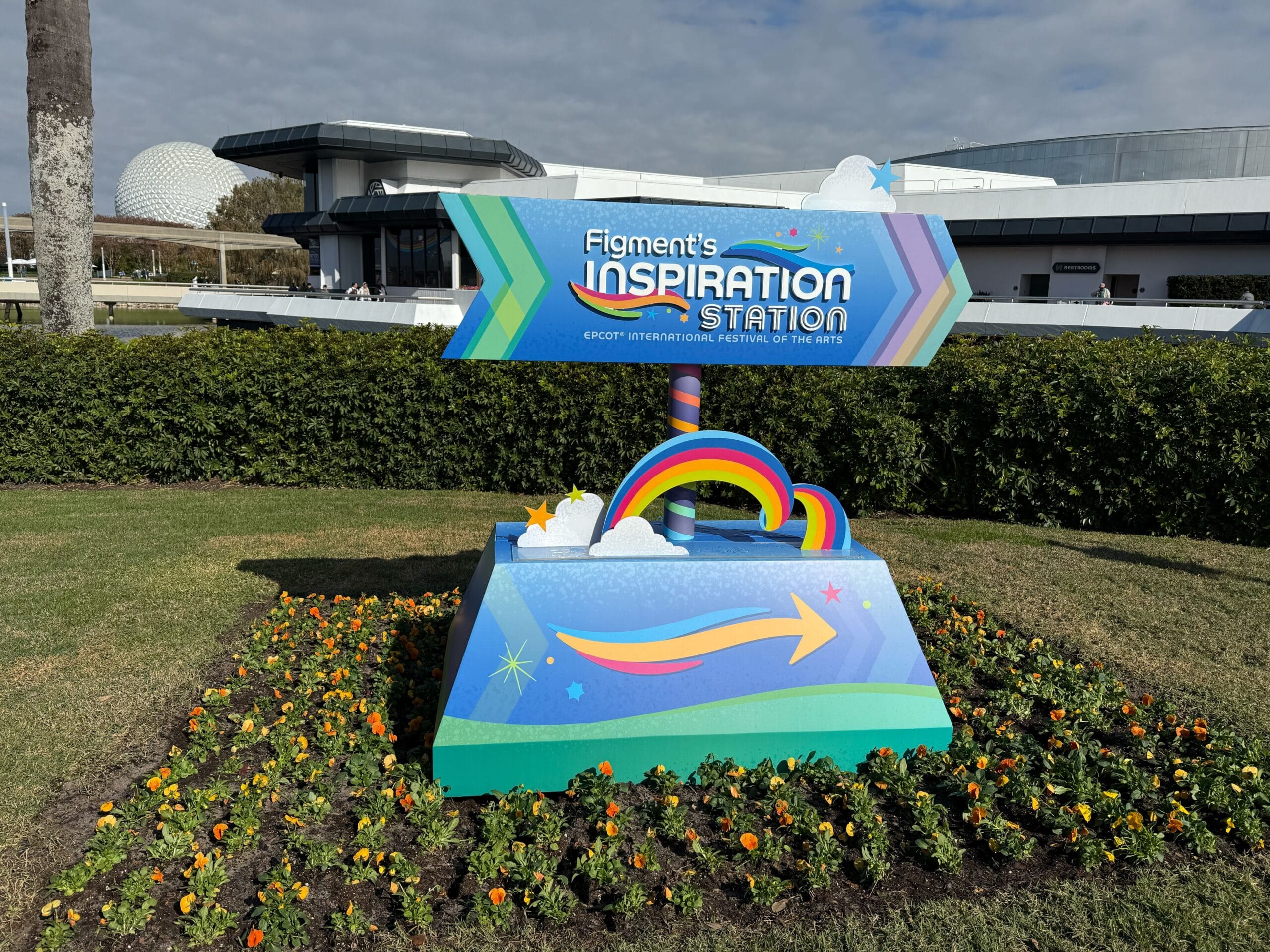The vibrant sign for "Figment's Inspiration Station" at Epcot, surrounded by rainbows and clouds, proudly stands amid a landscaped area adorned with flowers, perfectly capturing the spirit of the Festival of the Arts.