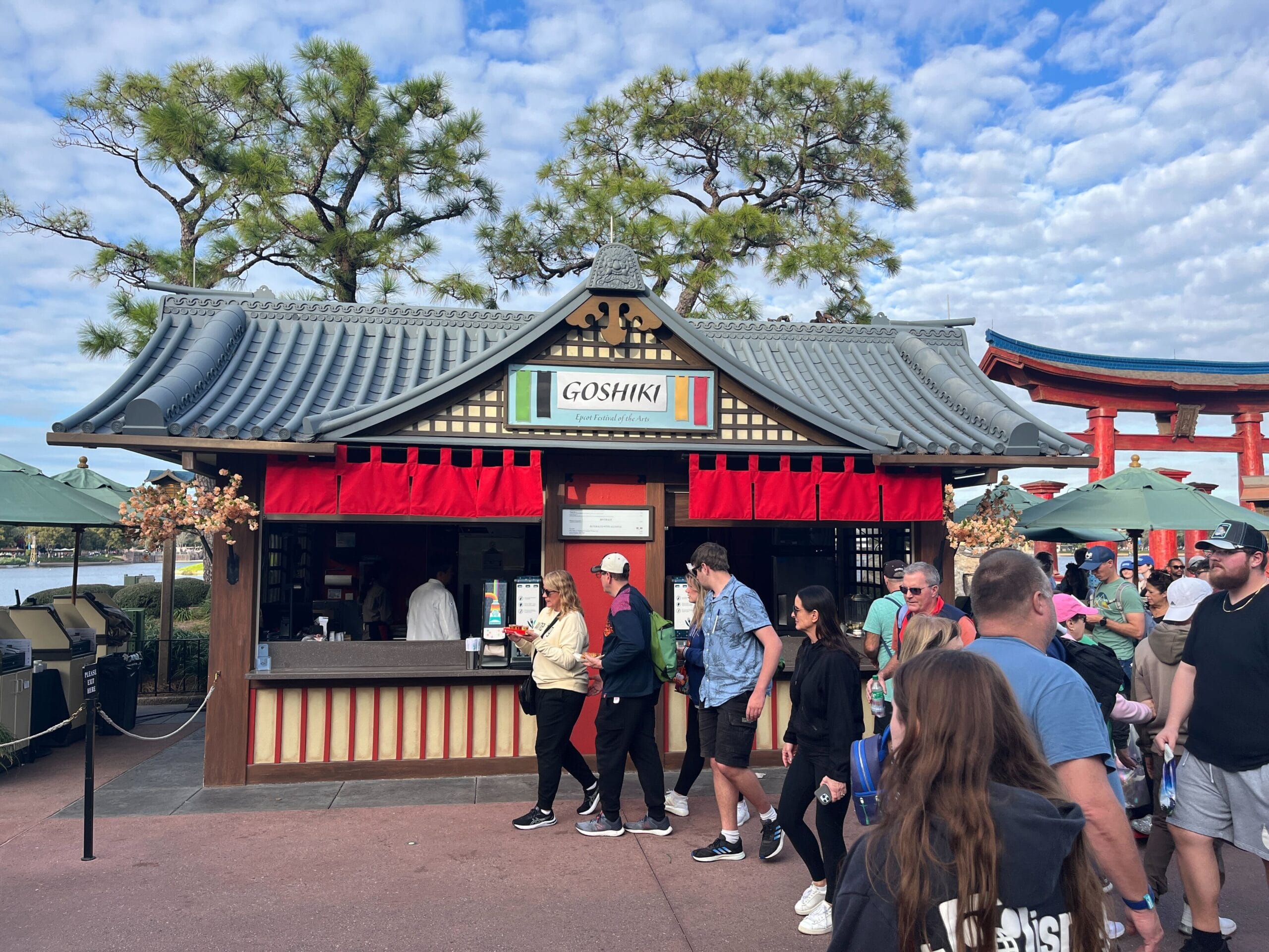 Amid the vibrant Festival of the Arts, people stroll past a Japanese-style food kiosk named "Goshiki," adorned with a traditional roof and red curtains, nestled against a backdrop of trees and a torii gate under the partly cloudy sky.