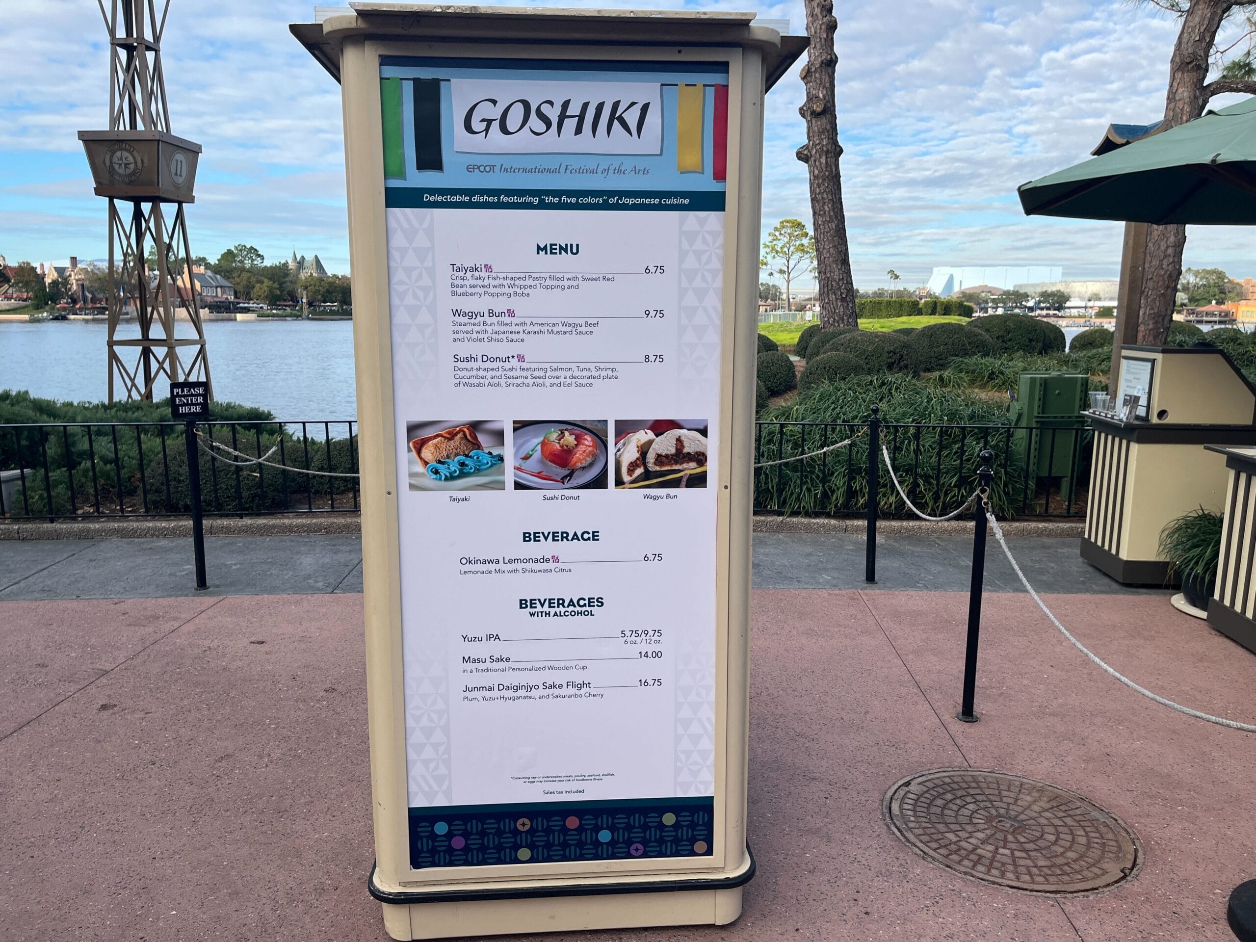 The outdoor menu board for Goshiki restaurant showcases delightful options like Taiyaki, Sushi Donut, and refreshing beverages. Set against a scenic waterfront view with trees gently swaying in the background, it perfectly complements the vibrant atmosphere of the Festival of the Arts.
