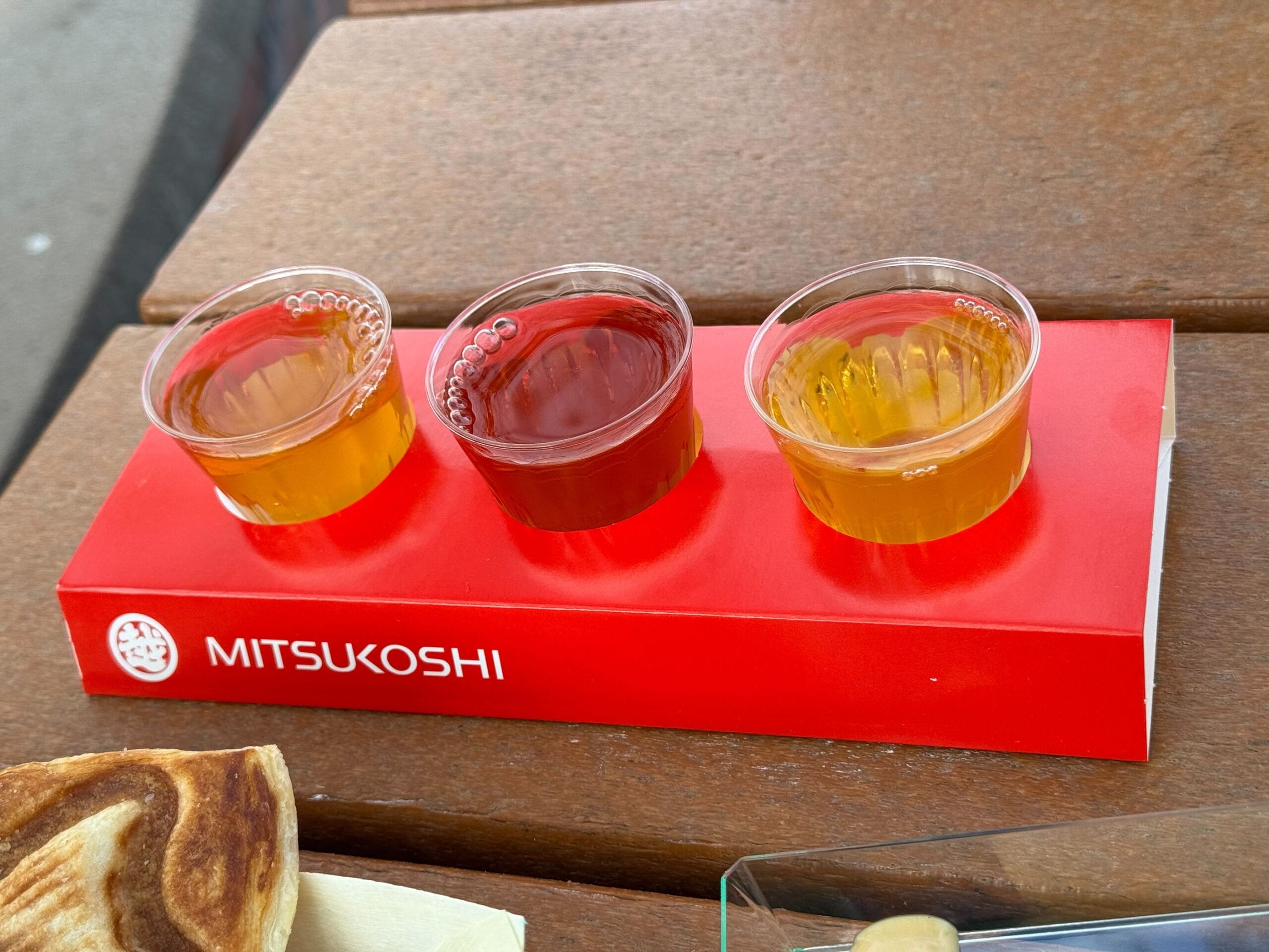 Three small cups of different colored liquid, reminiscent of the vibrant palette at the Festival of the Arts, are placed on a red Mitsukoshi branded tray resting on a wooden surface.