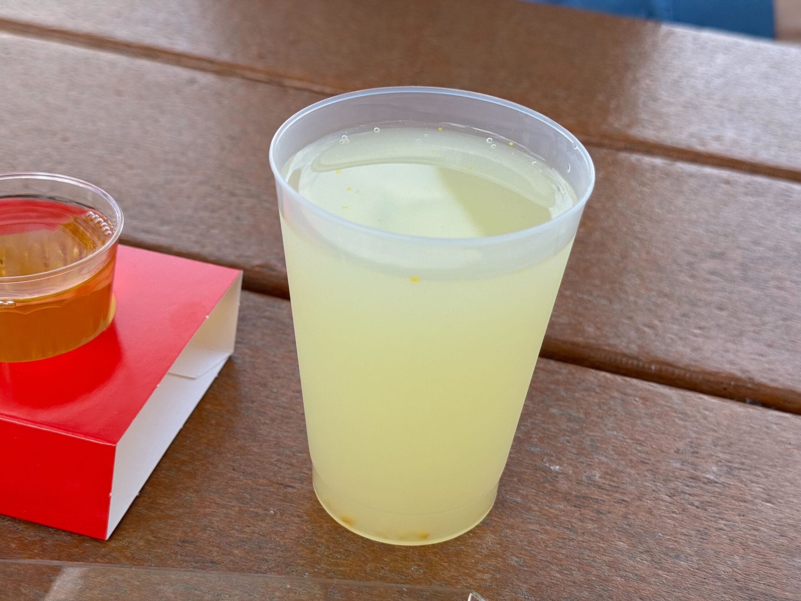 A plastic cup filled with lemonade sits on a wooden table next to a red box and a small cup with brown liquid, embodying the vibrant spirit of the Festival of the Arts.