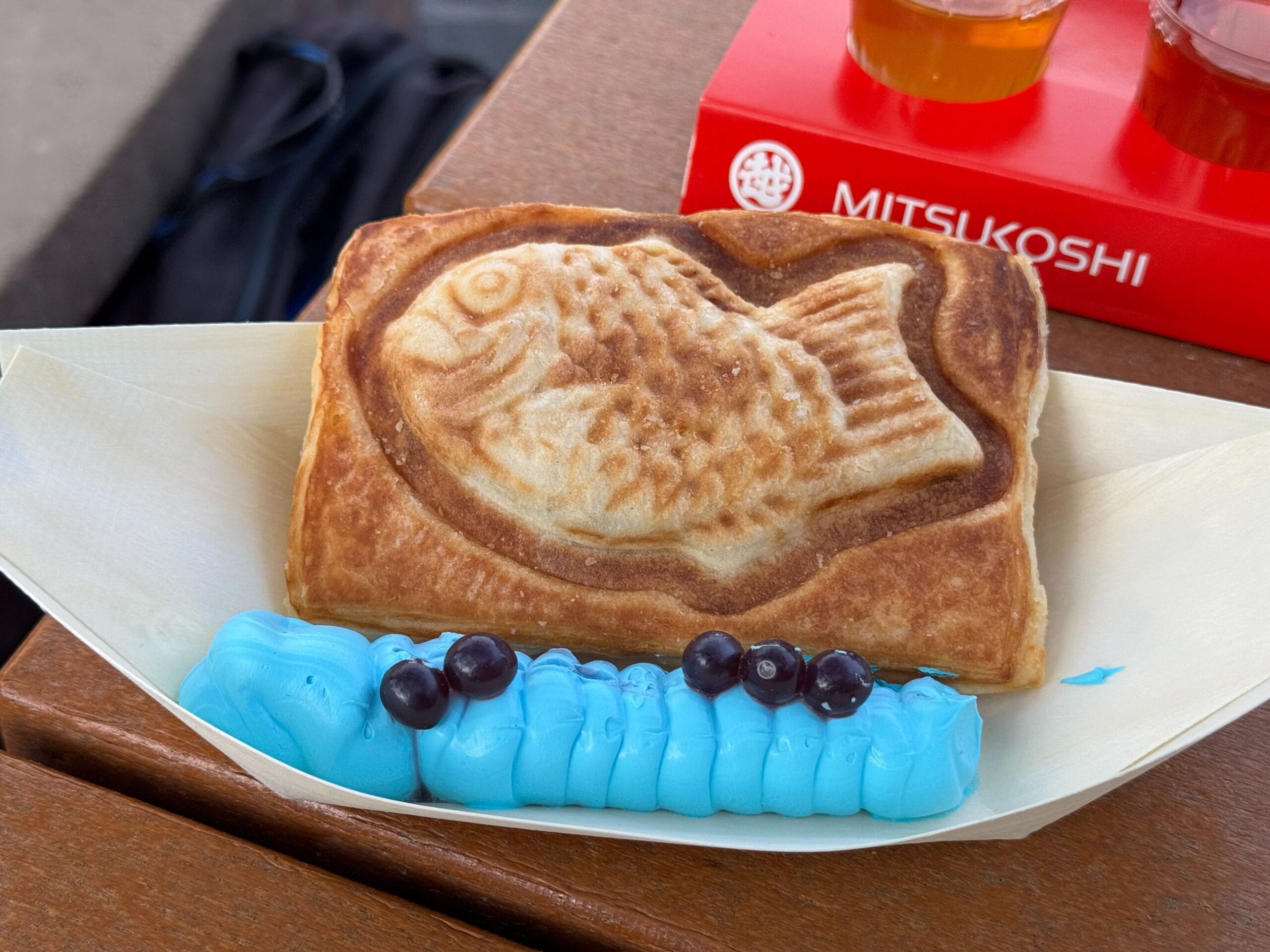 A fish-shaped pastry adorned with blue frosting and small round toppings rests on a paper tray, capturing the vibrant spirit of the Festival of the Arts. It sits beside a red box embossed with "MITSUKOSHI," creating a delightful tableau of color and artistry.