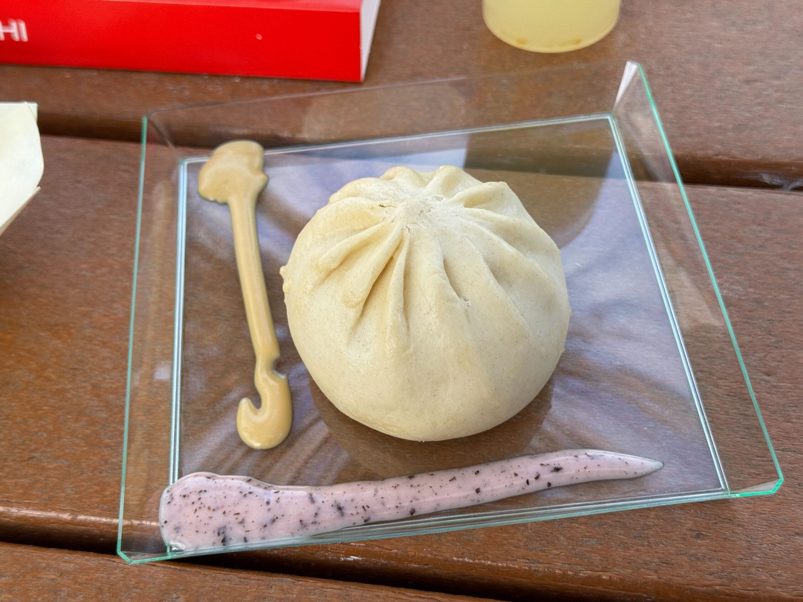 A steamed bun perched on a glass plate, its presentation worthy of the Festival of the Arts, with two sauces: one light brown in a graceful curve and another speckled white in a playful zigzag.