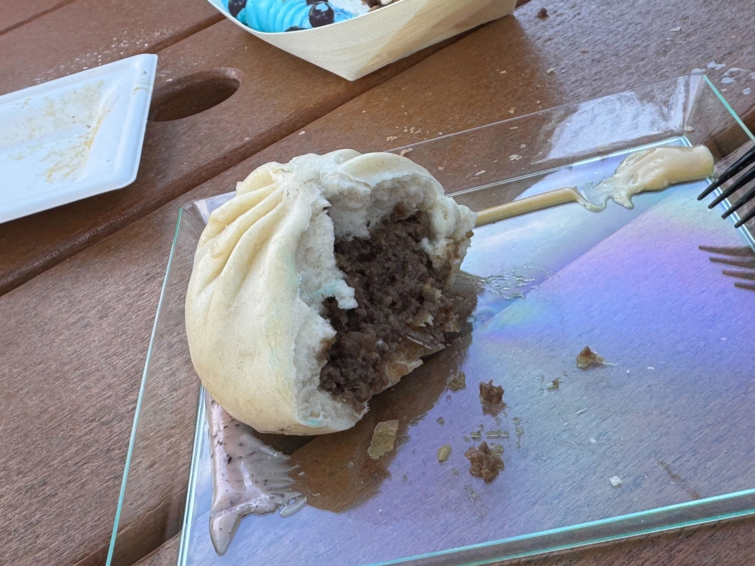 A half-eaten steamed bun with savory meat filling rests on a glass plate, surrounded by scattered crumbs. In the background, another dish peeks into view, invoking the creative spirit of a Festival of the Arts feast.