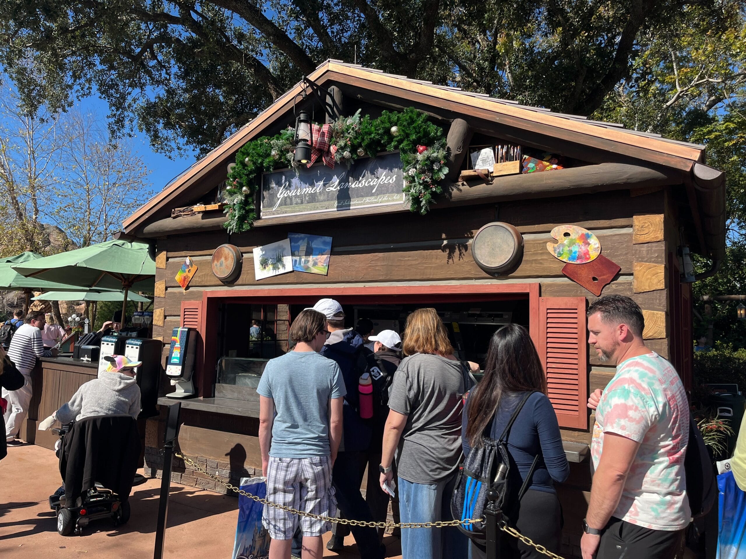 People wait in line at a wooden food kiosk, adorned with greenery and postcards, soaking up the vibrant atmosphere of the Festival of the Arts.