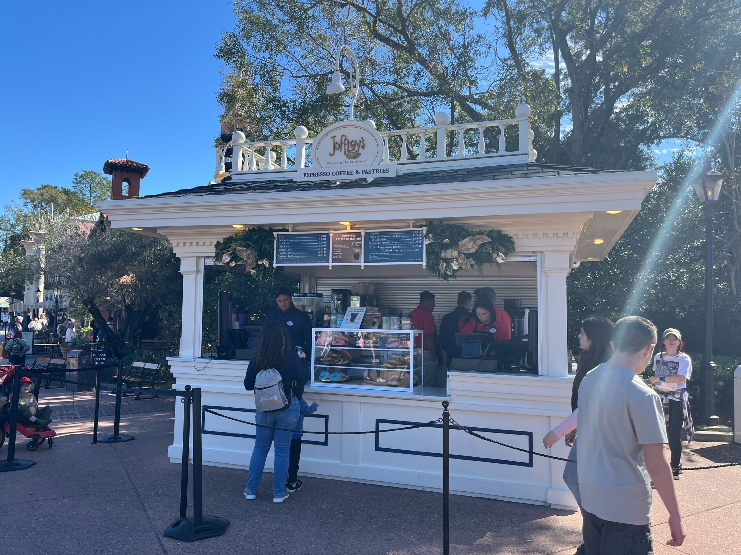 Outdoor coffee and pastry kiosk with the "Joffrey's" sign, nestled amid a vibrant Festival of the Arts. Surrounded by a few people enjoying the ambiance, trees stretch into a clear blue sky above.