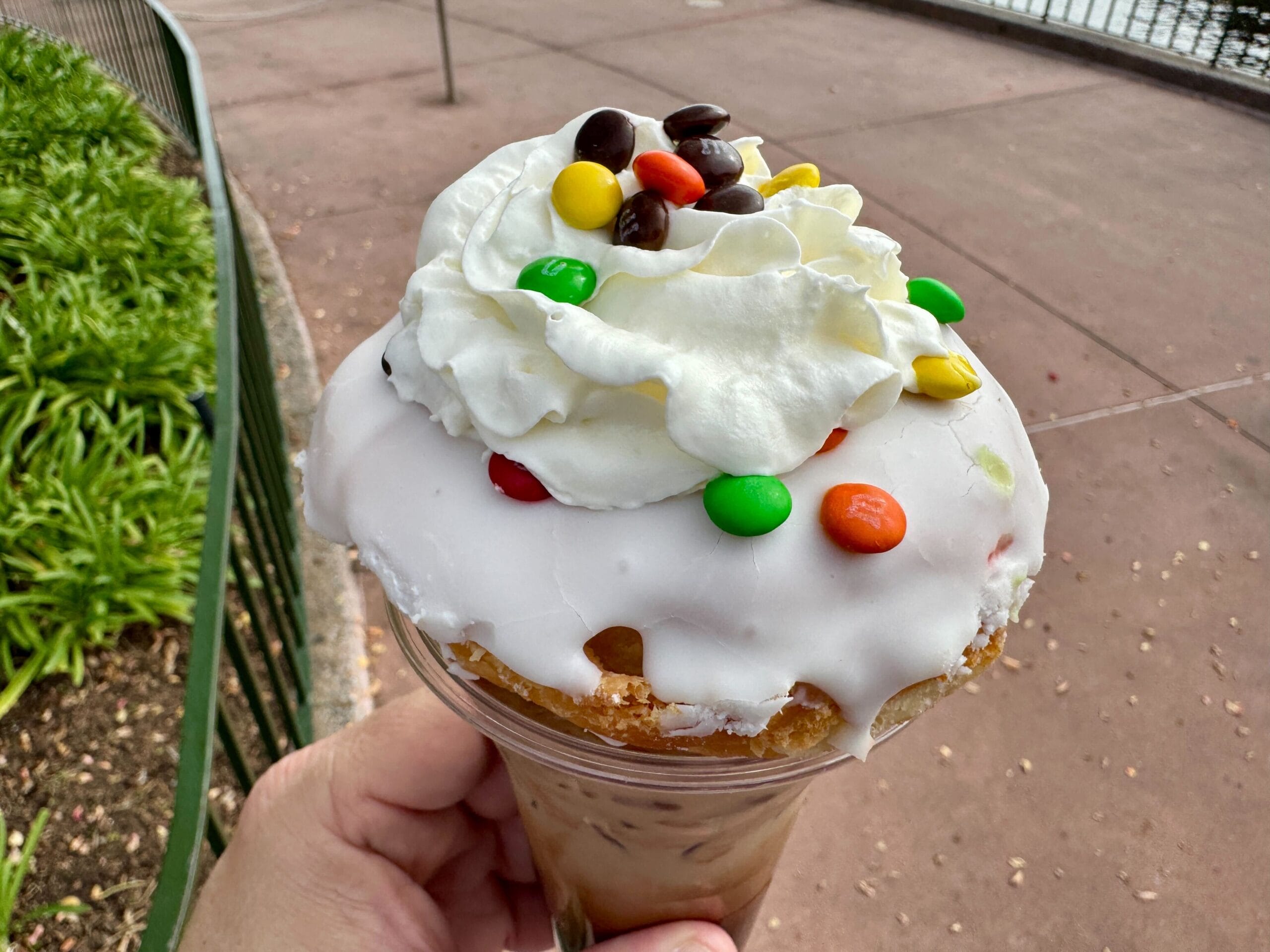 Ice cream cone topped with whipped cream and colorful candy-coated chocolates, held outdoors near a railing.