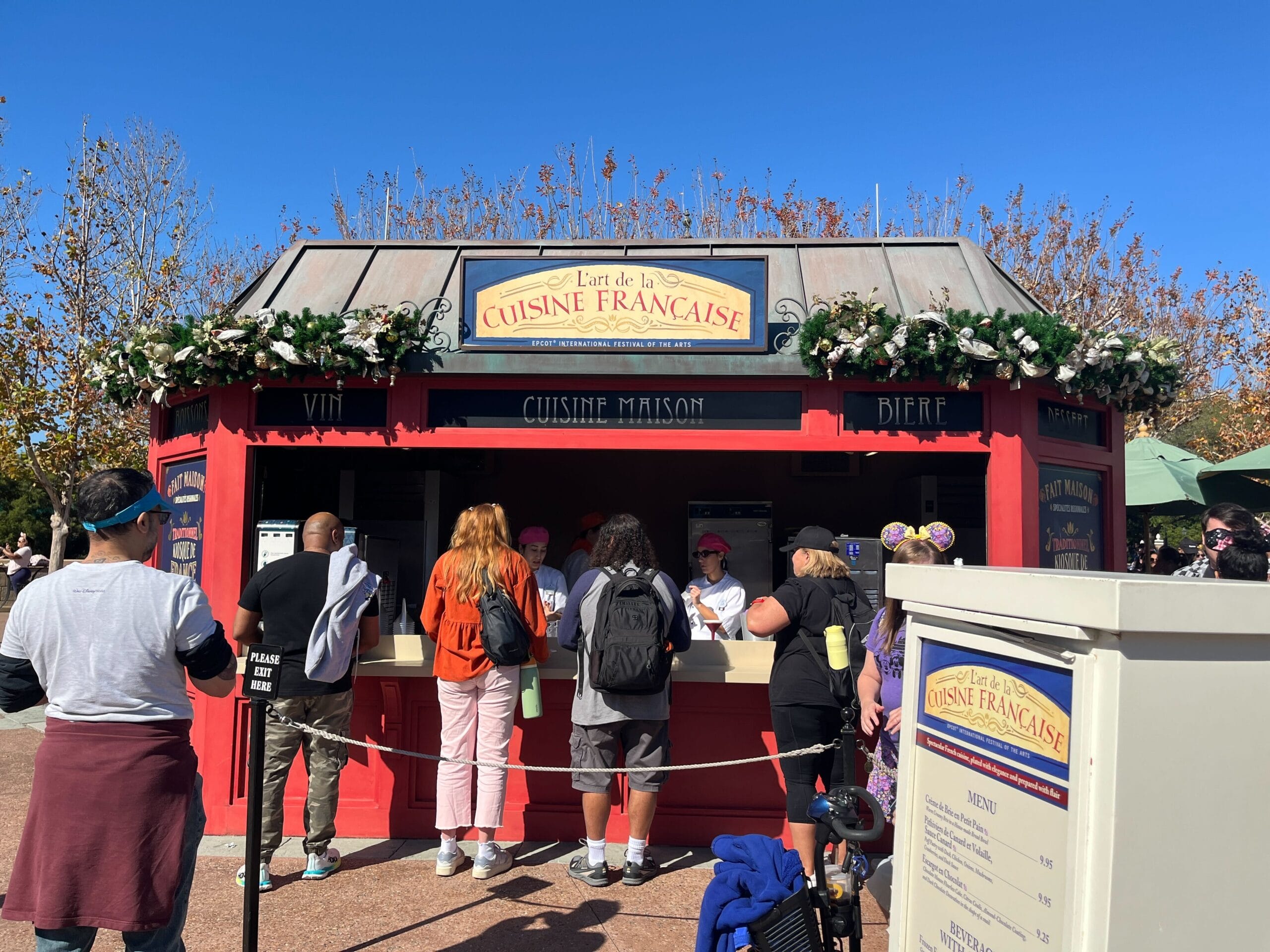 People wait in line at a red booth labeled "Cuisine Française," adorned with greenery and flowers, at the lively Festival of the Arts. With a menu board displayed, this charming spot serves delicious food and drinks.