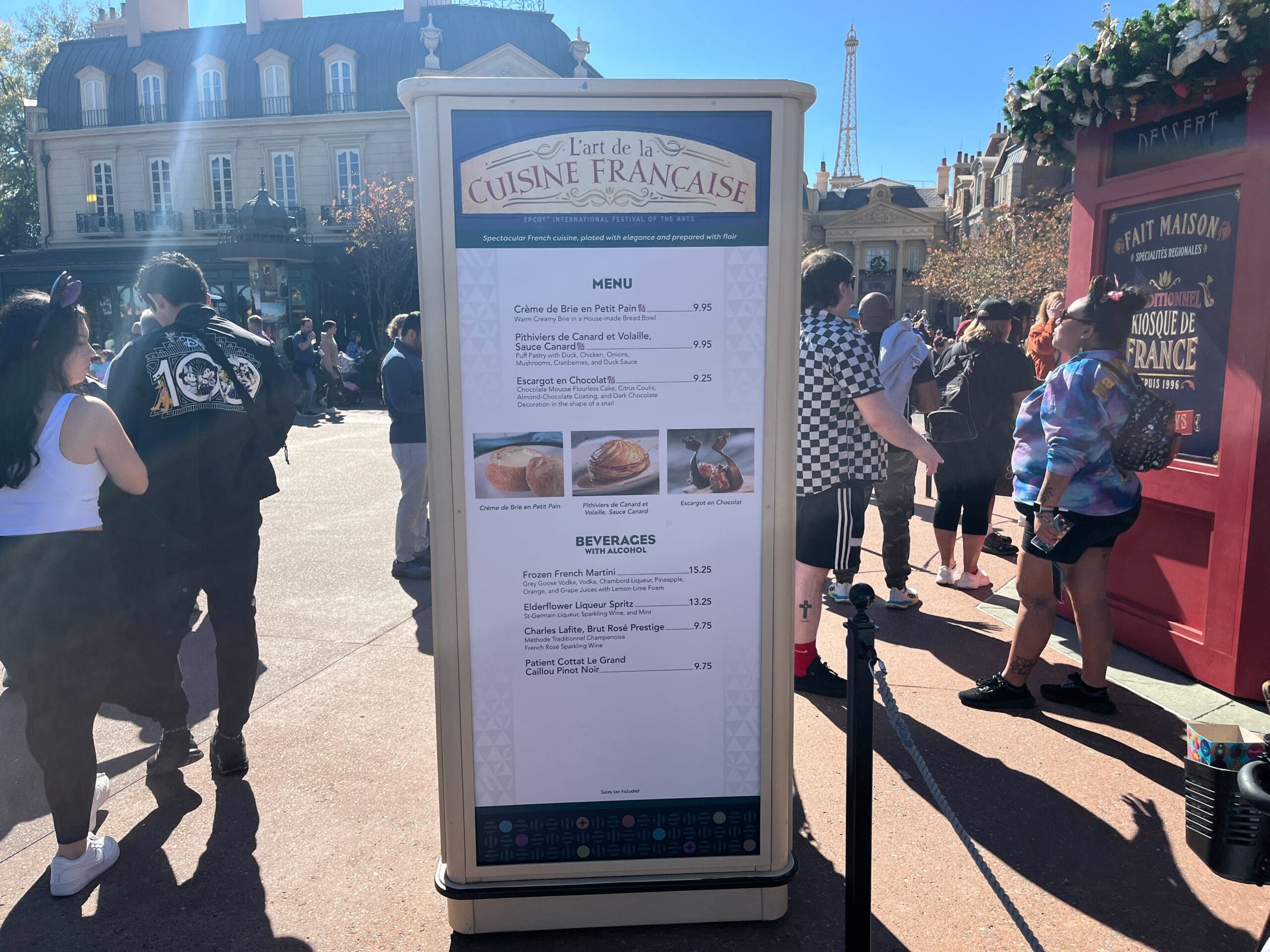 A sign titled "L'art de la Cuisine Française" displays a menu with items and prices. Celebrating the Festival of the Arts, people stand nearby, savoring the vibrant atmosphere as a building with elegant French architecture serves as an enchanting backdrop.