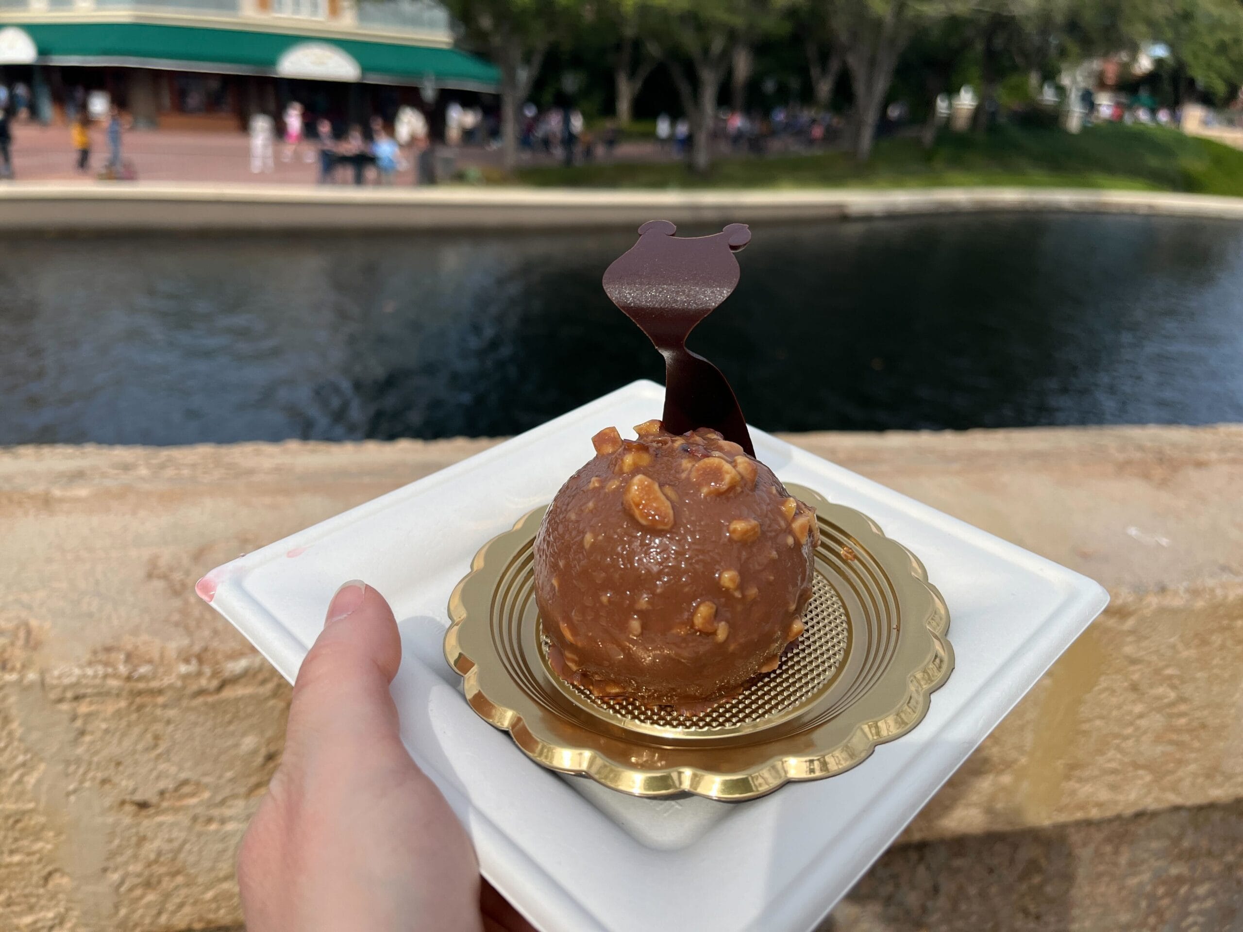 A hand holds a chocolate dessert adorned with nuts and a delicate chocolate decoration, resting on a gold and white plate. The scene captures the essence of the Festival of the Arts, set beside a tranquil canal lined with trees and framed by an elegant building in the background.