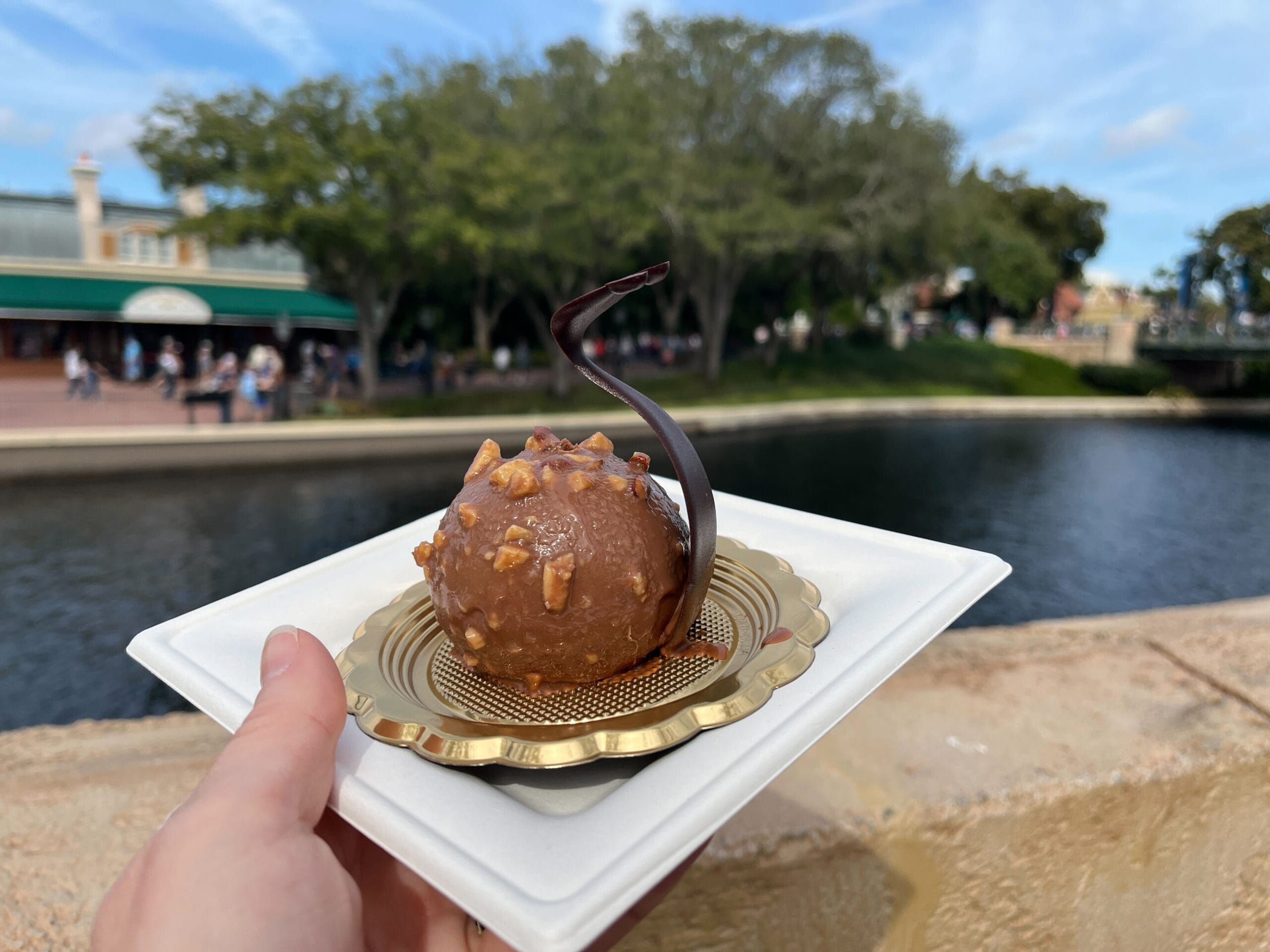 A hand holds a plate with a chocolate dessert, adorned with a curved chocolate piece and nuts, set against the enchanting backdrop of a river and trees, reminiscent of the Festival of the Arts ambiance.