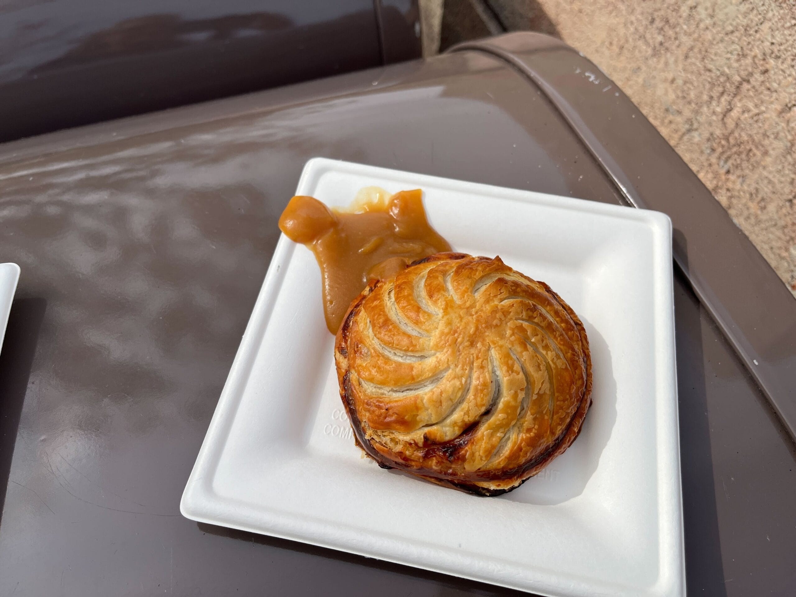 A pastry with a decorative swirl pattern on top, echoing the creative essence of the Festival of the Arts, served on a white tray with caramel or sauce beside it, all beautifully arranged on a brown surface.