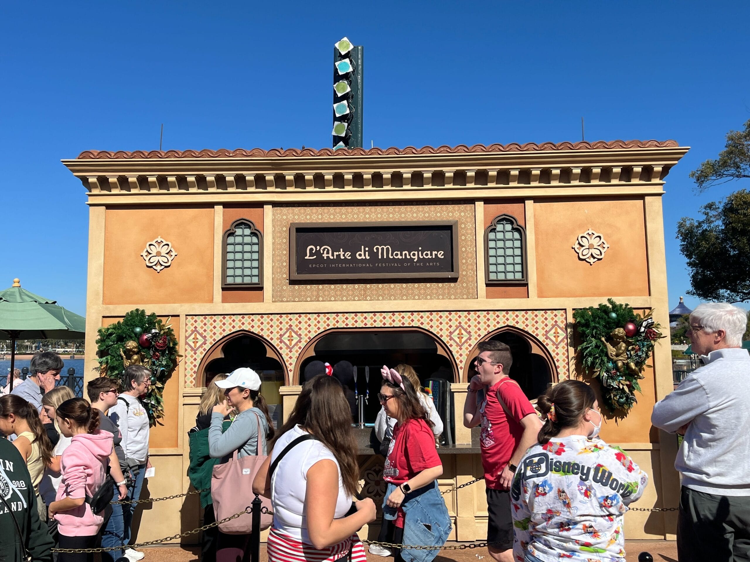 People standing in line outside a building labeled "L'Arte di Mangiare," adorned with festive decorations for the Festival of the Arts, under a clear blue sky.