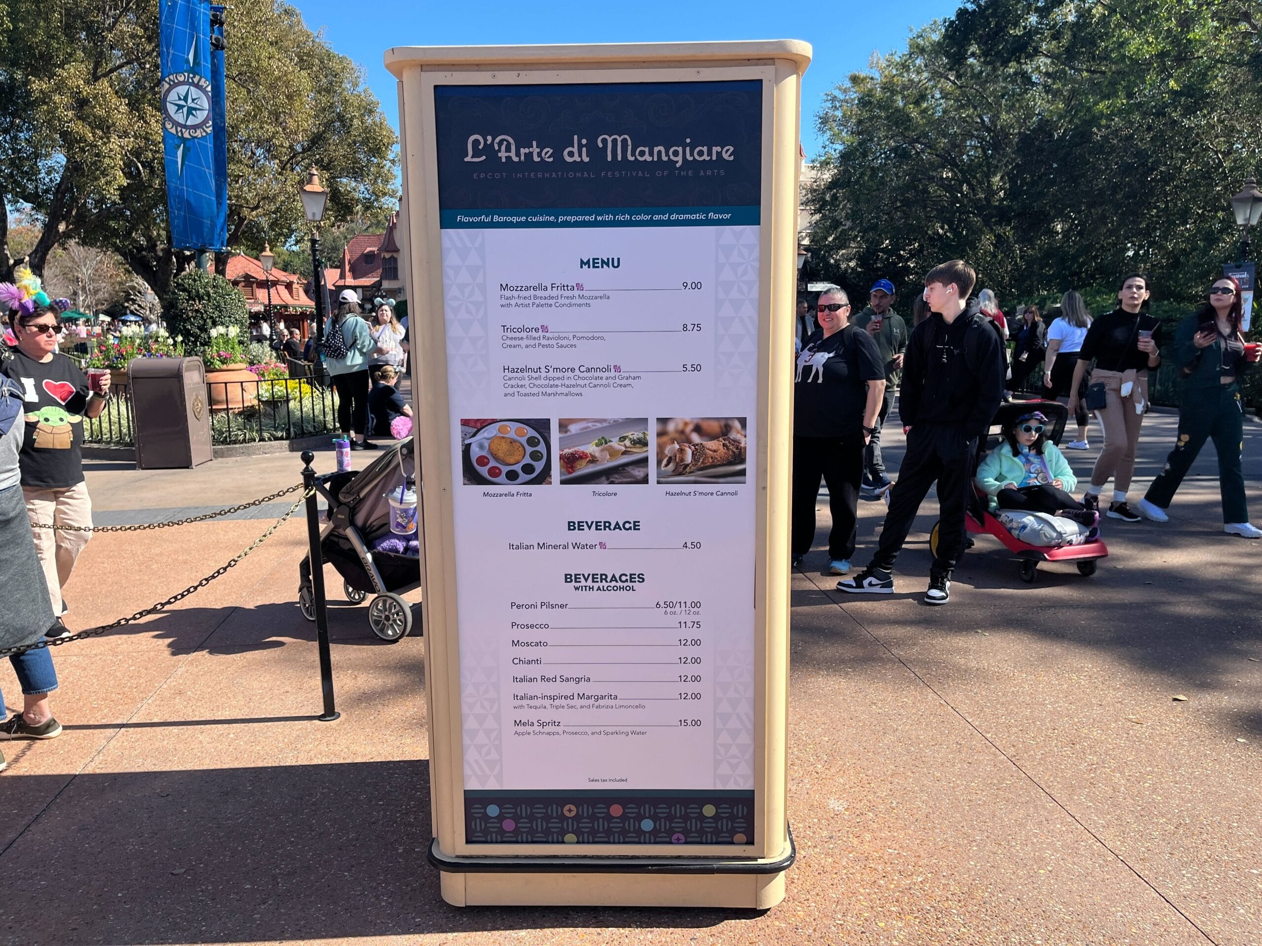 The menu board at "L’Arte di Mangiare," adorned with artistic flair for the Festival of the Arts, displays food and beverage options. Nearby, people stroll and stand, with a stroller subtly in the background.