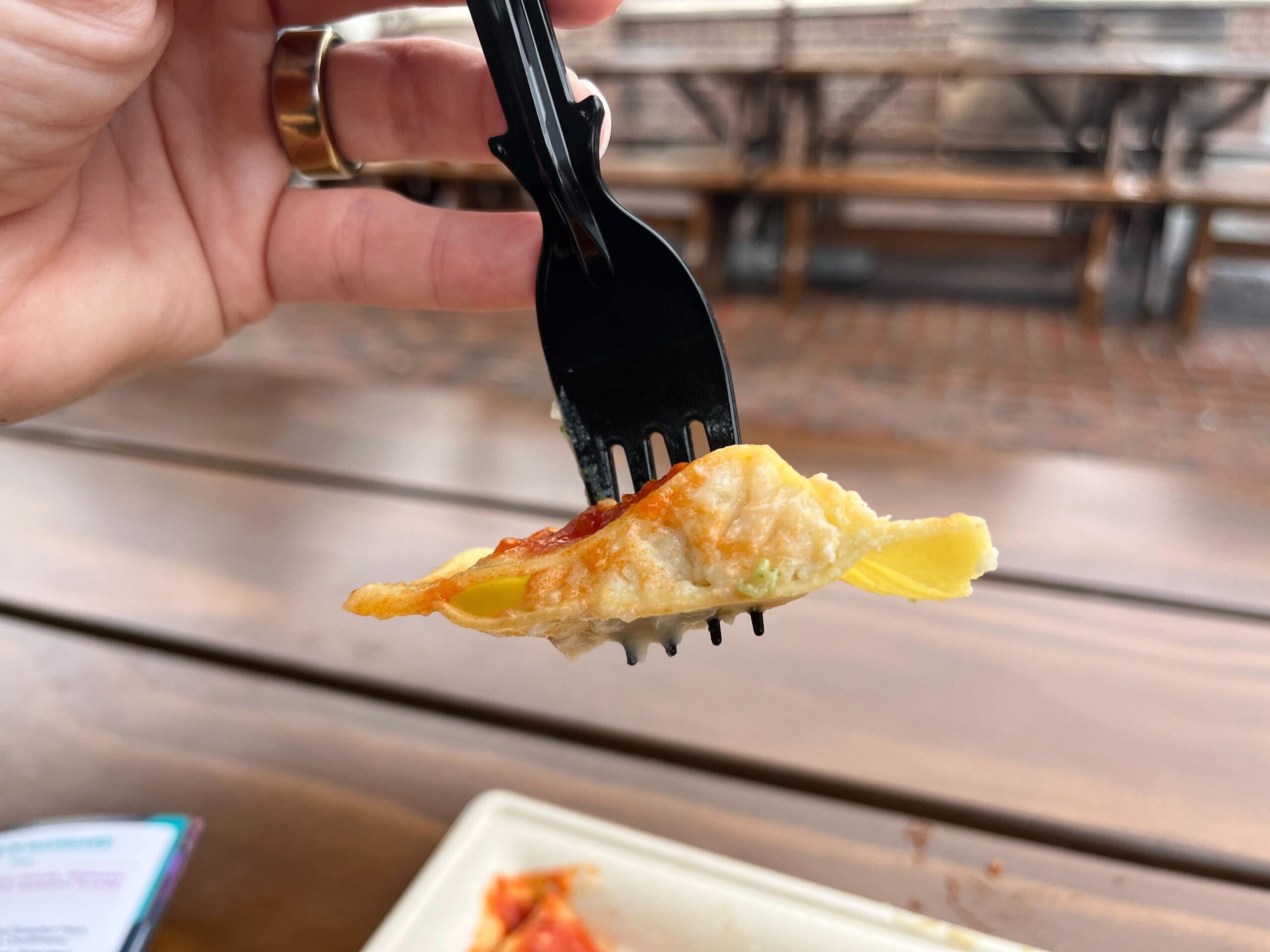 A hand gracefully holds a black fork with a bite of food, possibly a piece of omelette with vegetables, over an outdoor wooden table, embodying the culinary creativity celebrated at the Festival of the Arts.