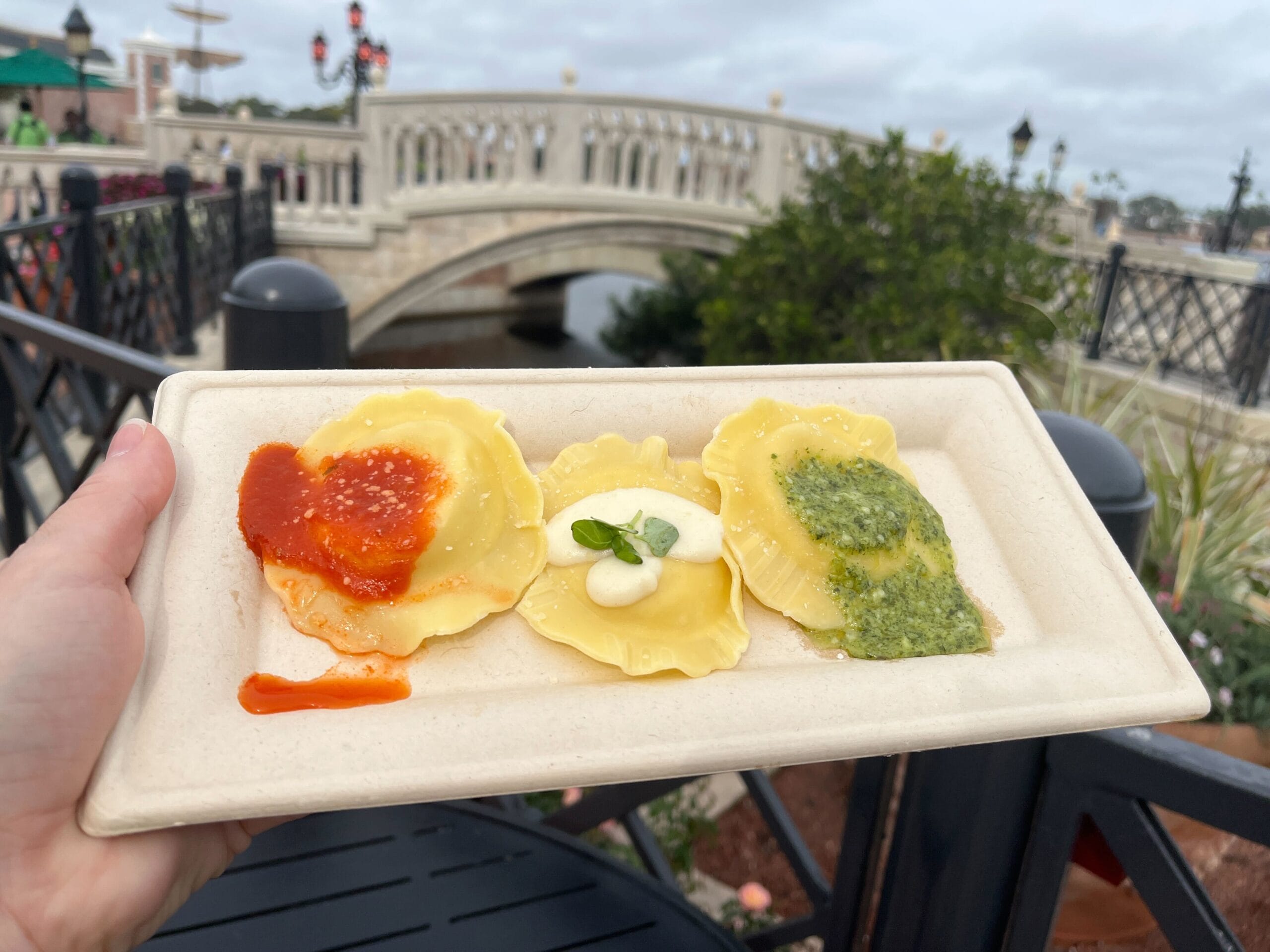 A hand presents a plate with three ravioli, adorned with red sauce, cream, and green pesto, in a scene evocative of the Festival of the Arts. In the background, a stone bridge arches gracefully over a tranquil canal.