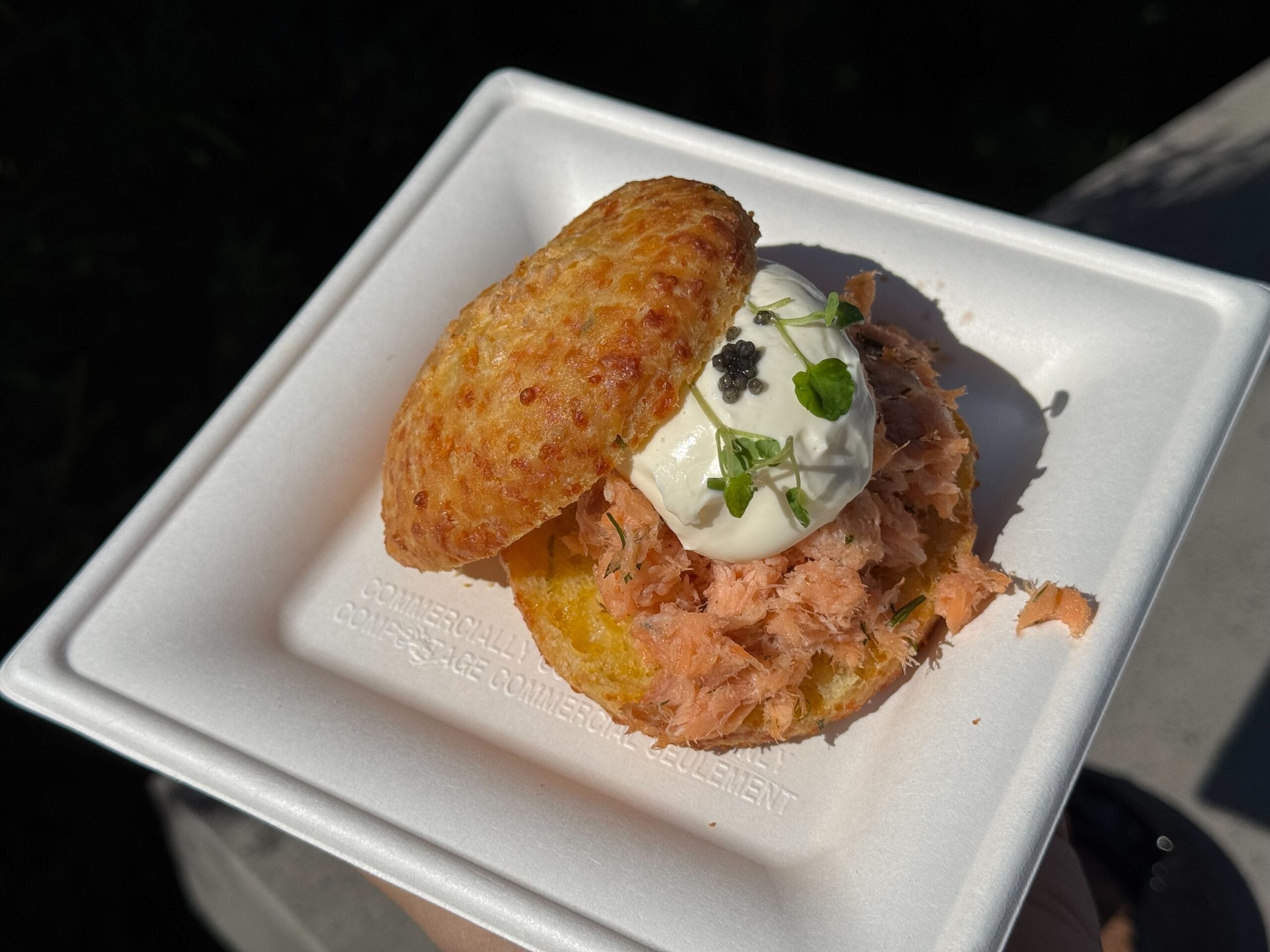 A delightful small sandwich featuring shredded salmon, a dollop of cream, and herbs, elegantly served on a biscuit in a white foam container. Perfect for savoring while exploring the Festival of the Arts.