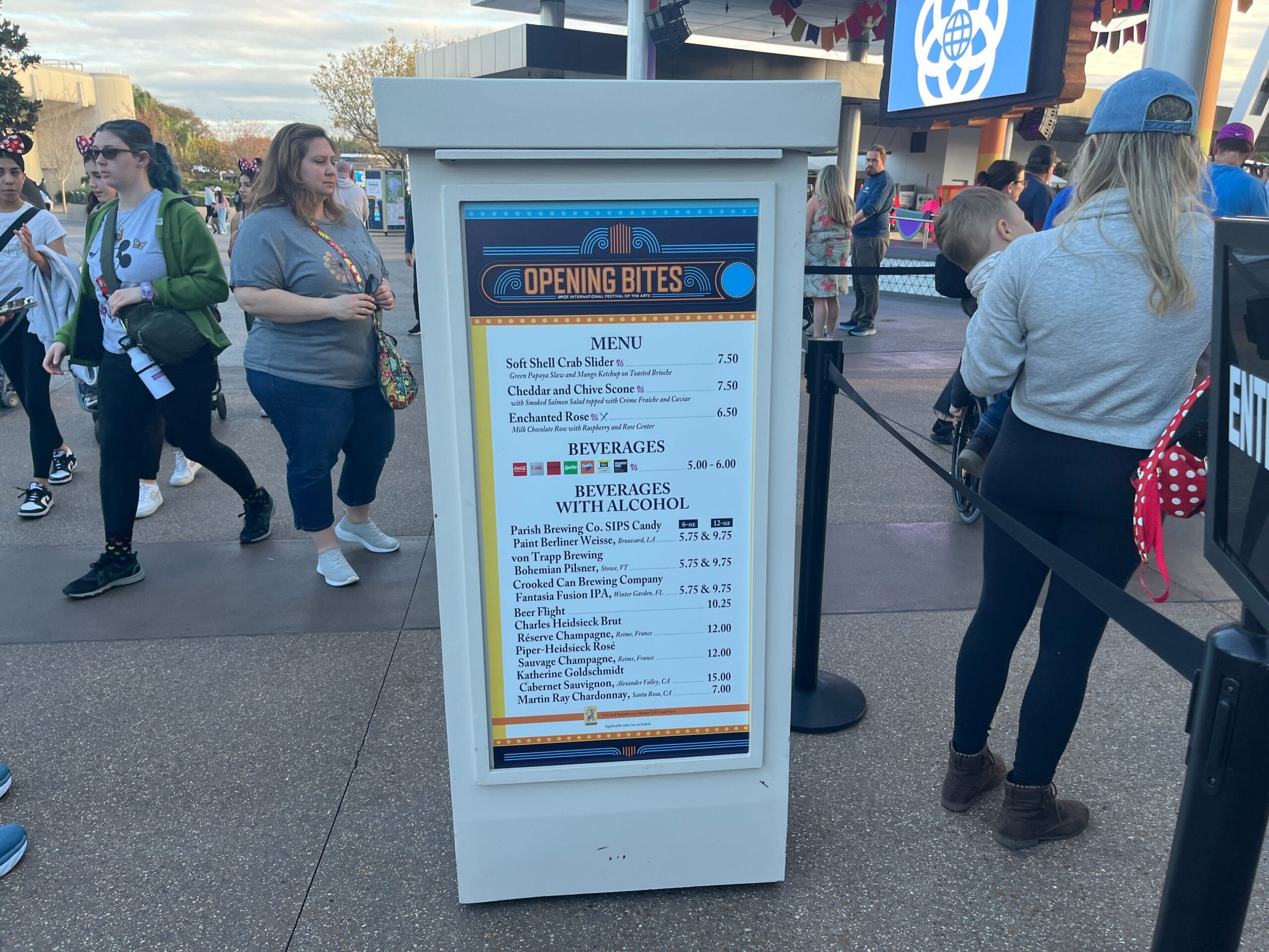 A menu sign for "Opening Bites" at the Festival of the Arts showcases delectable options like soft shell crab sliders and cocktails. In the background, pedestrians wander near a vibrant theme park entrance, adding to the festive atmosphere.