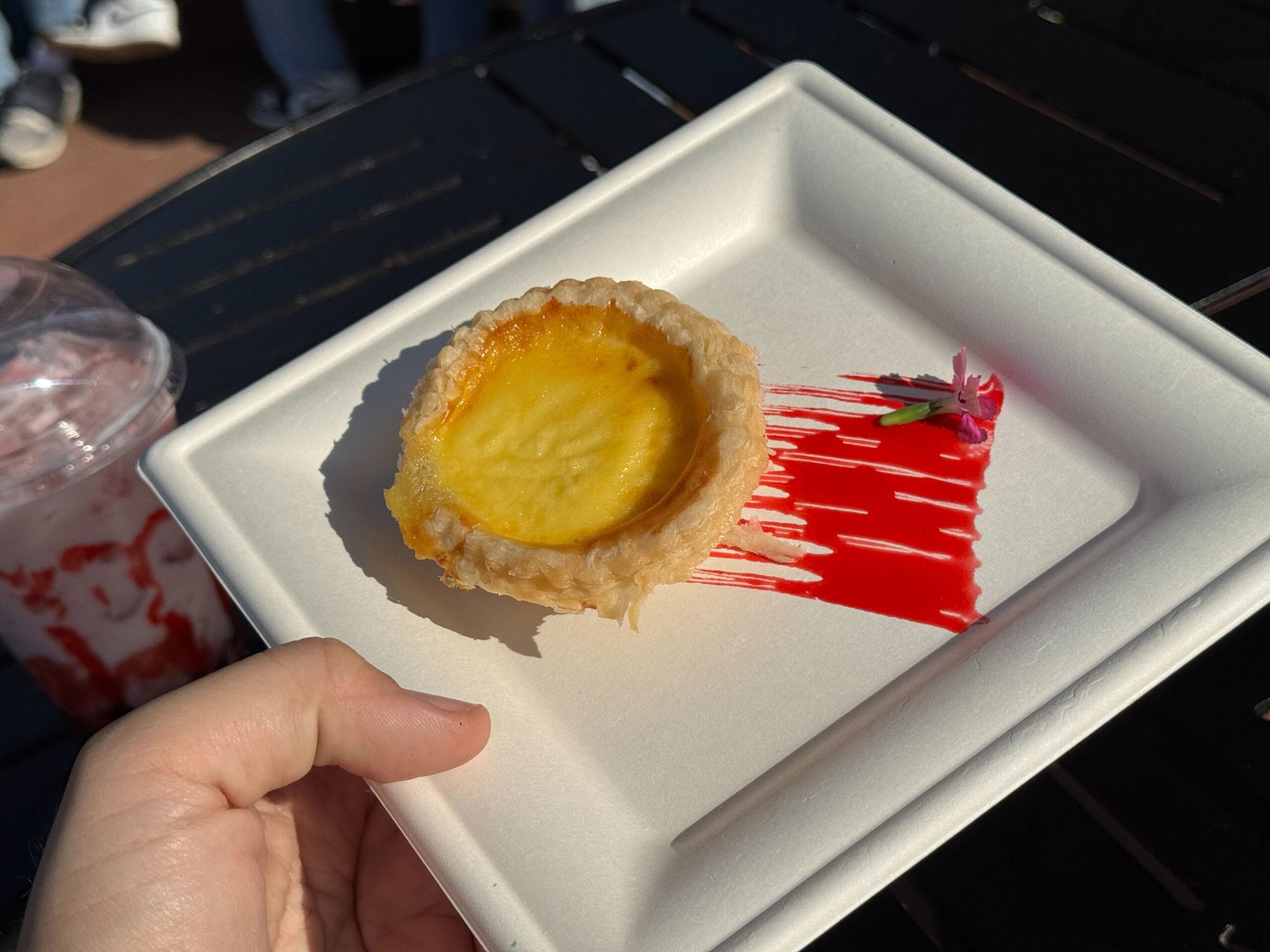 A hand holds a plate with a small tart and red sauce streak, reminiscent of the Festival of the Arts. A pink flower and green garnish are beside the tart, adding artistic flair, while a drink is partially visible in the background on a dark table.