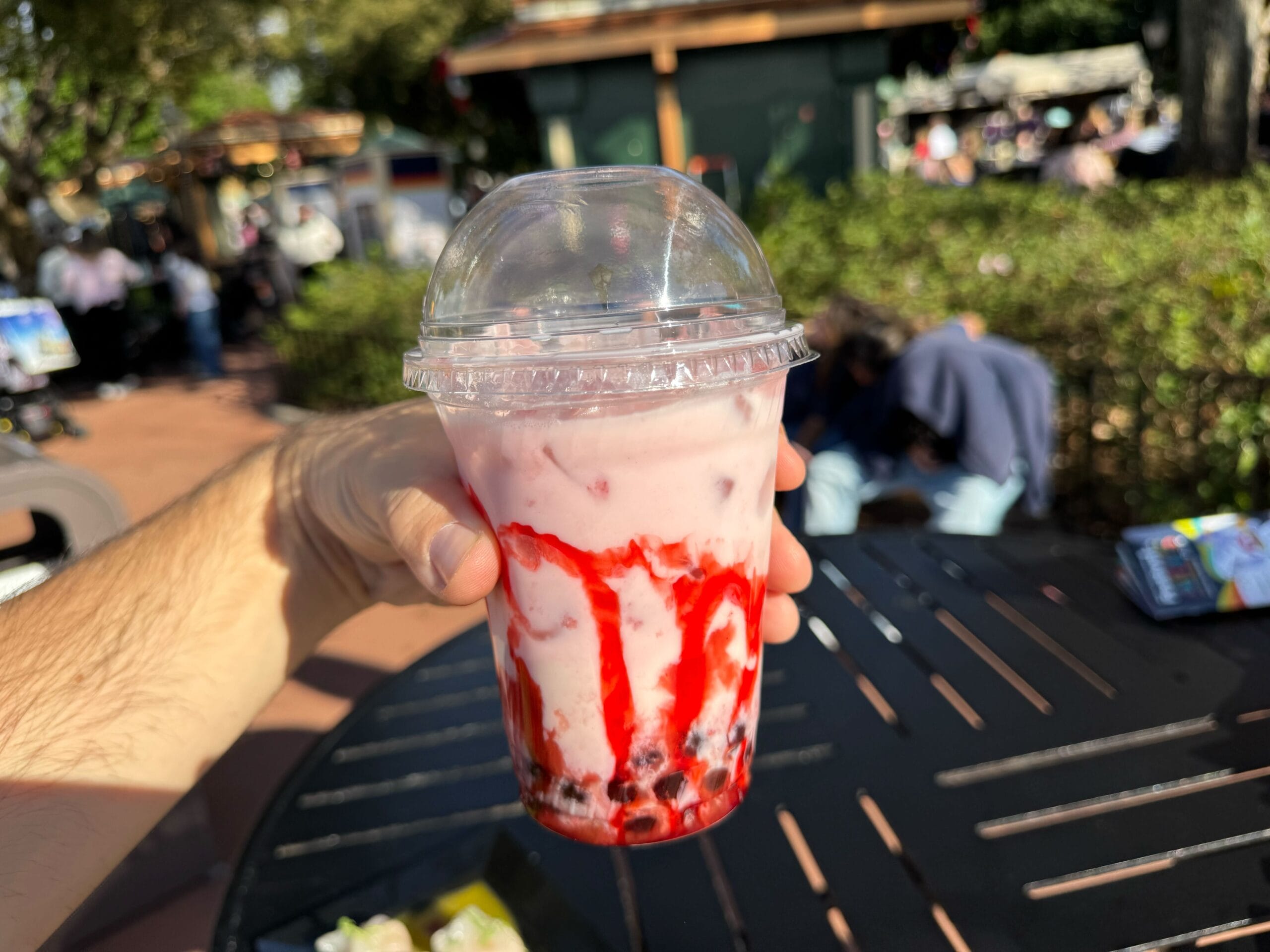 A hand holds a plastic cup containing a pink and red drink with a domed lid, capturing the vibrant spirit of the Festival of the Arts on a sunny day.