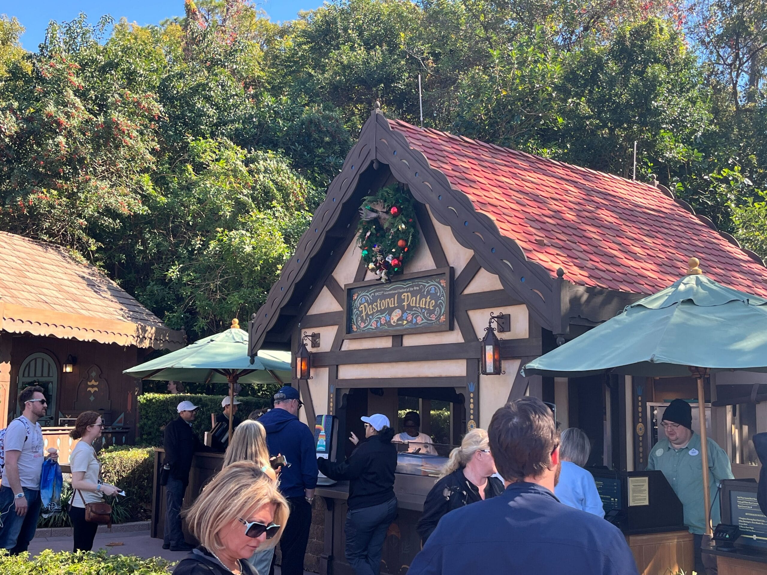 People standing in line at a small wooden stall with a red roof and wreath decoration, surrounded by trees, eagerly await their turn at the Festival of the Arts.