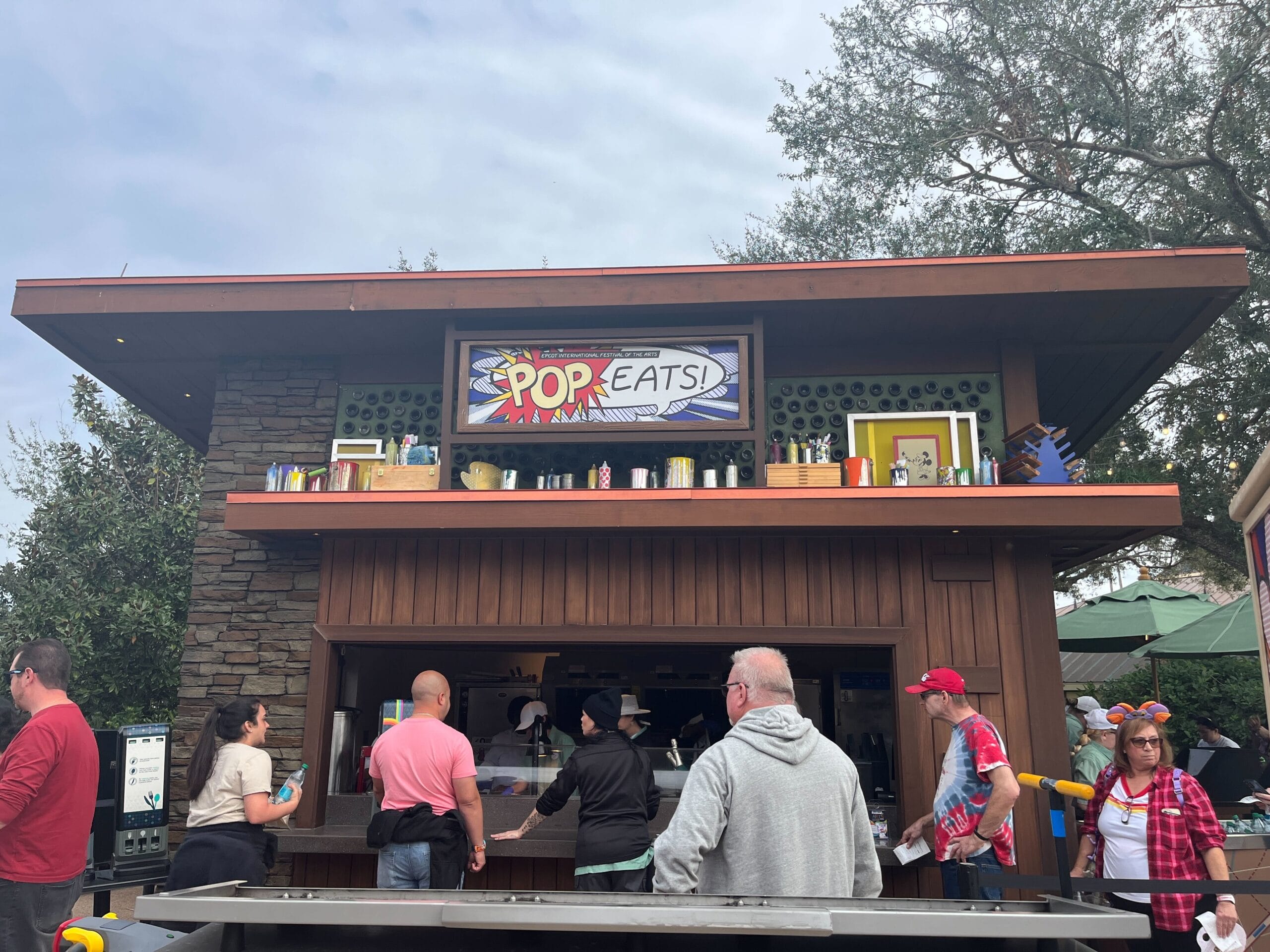 Pop Eats! is a small food kiosk bustling with patrons in line, set against the vibrant backdrop of the Festival of the Arts. The counter is staffed by enthusiastic employees, and art-themed decorations adorn the roof edge, while trees gently sway in the background.