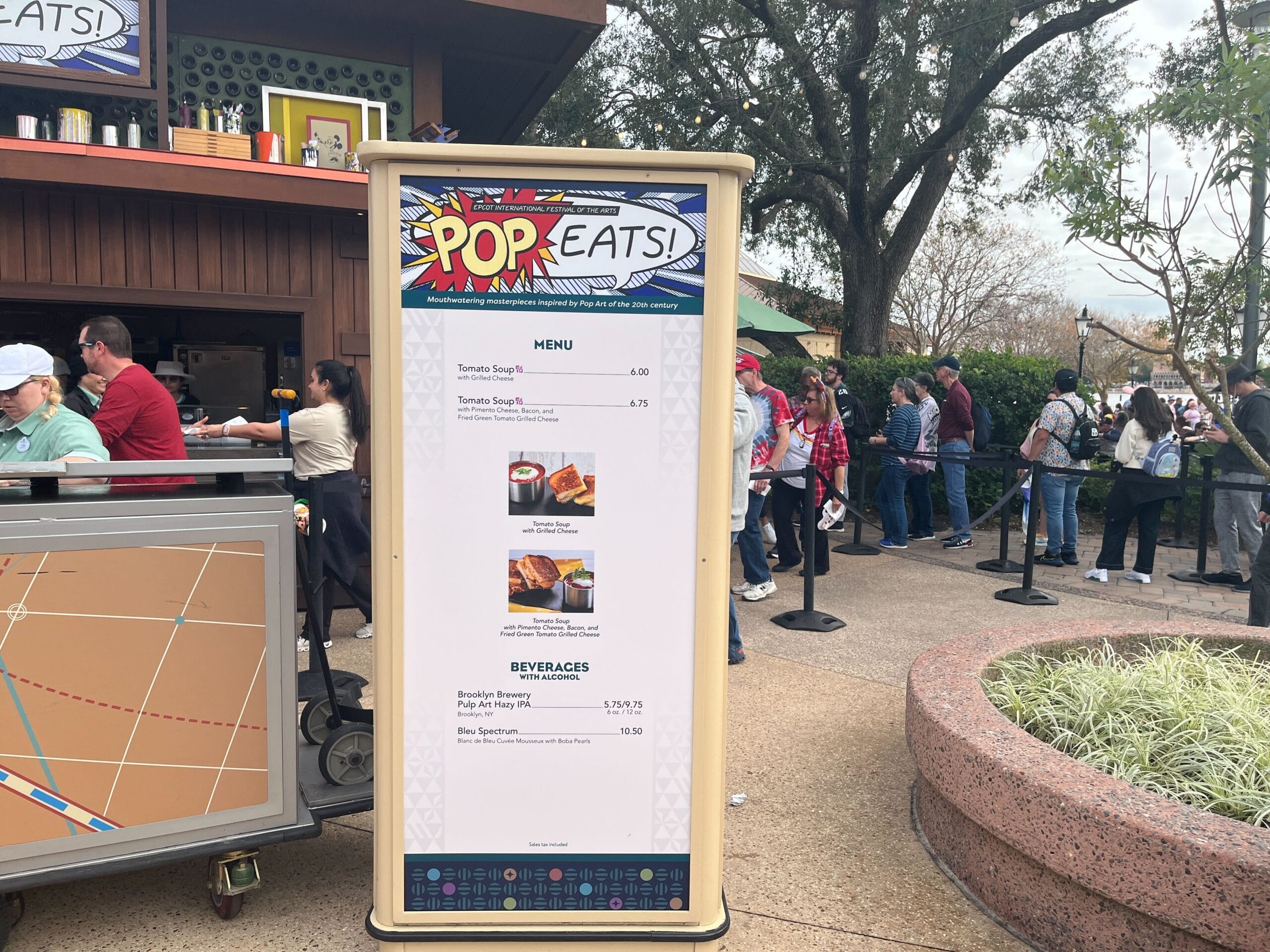 Sign displaying the Festival of the Arts menu at Pop Eats! booth with tomato soup and grilled cheese options; line of people in background.
