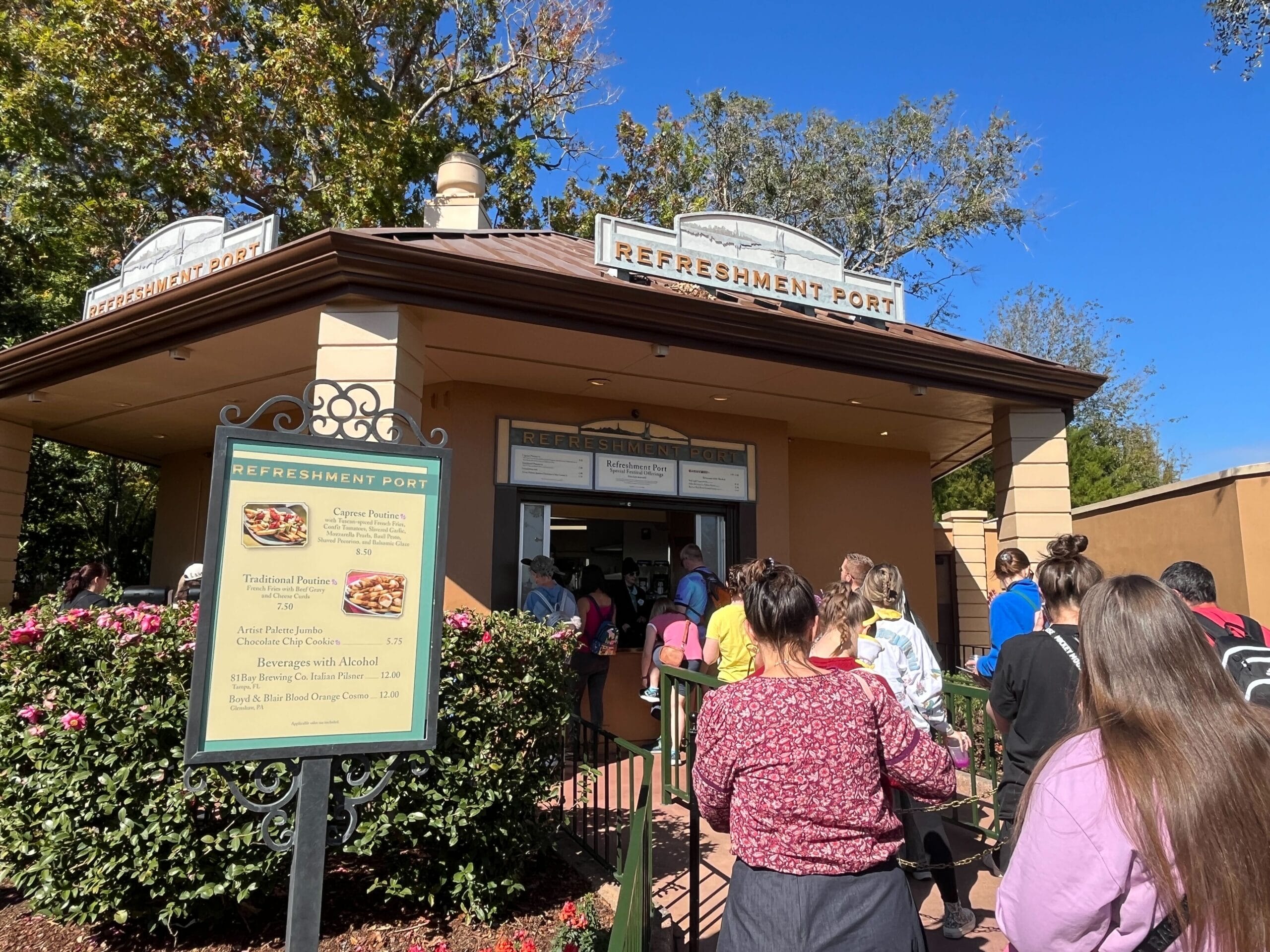 People standing in line at a refreshment port during the Festival of the Arts on a sunny day, with a menu board displaying food options at the entrance.