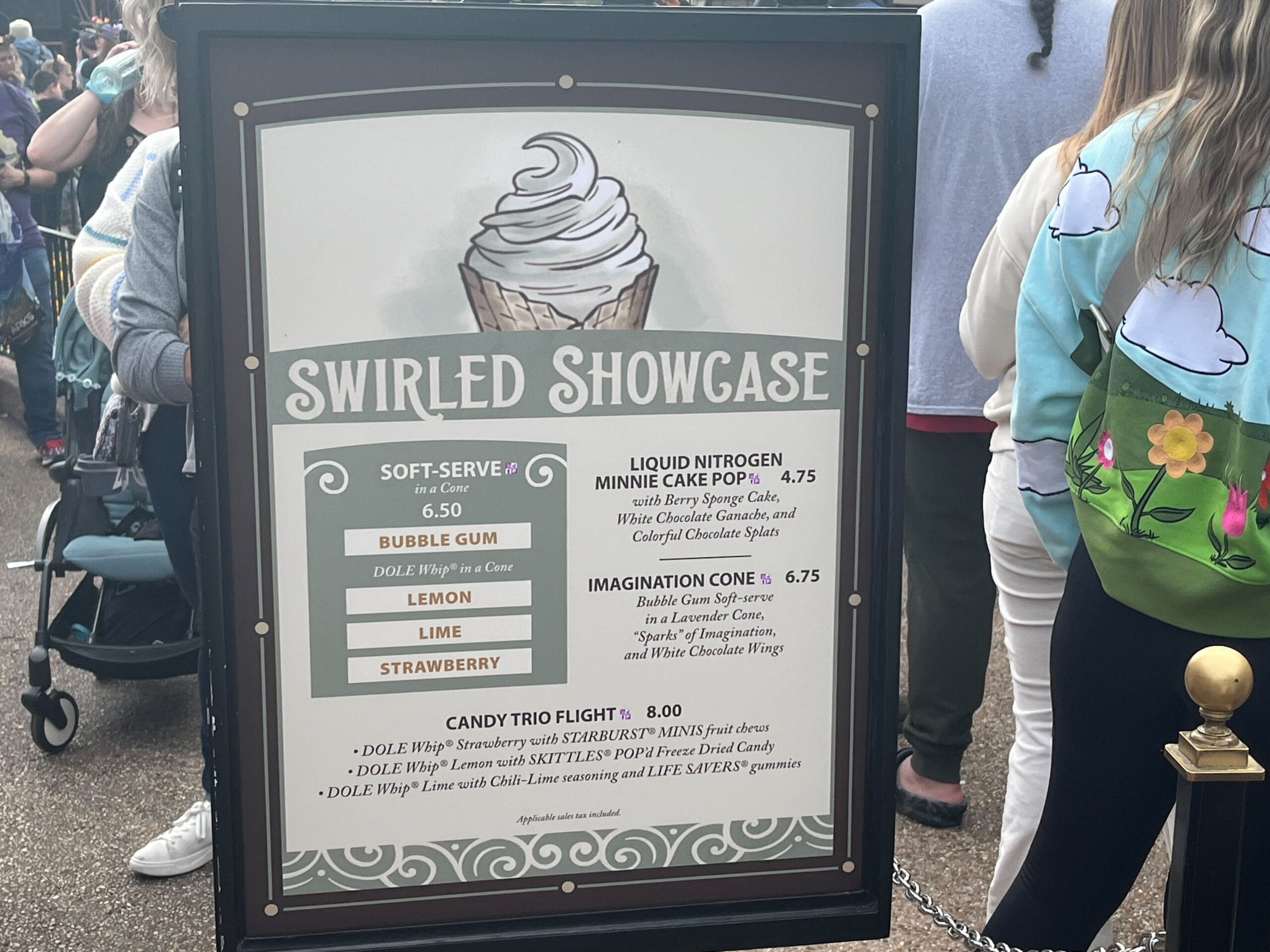 A sign highlights the Festival of the Arts' mouthwatering ice cream options, showcasing soft-serve flavors, liquid nitrogen cake pops, and candy trio flights, as a line of eager festival-goers waits in anticipation.