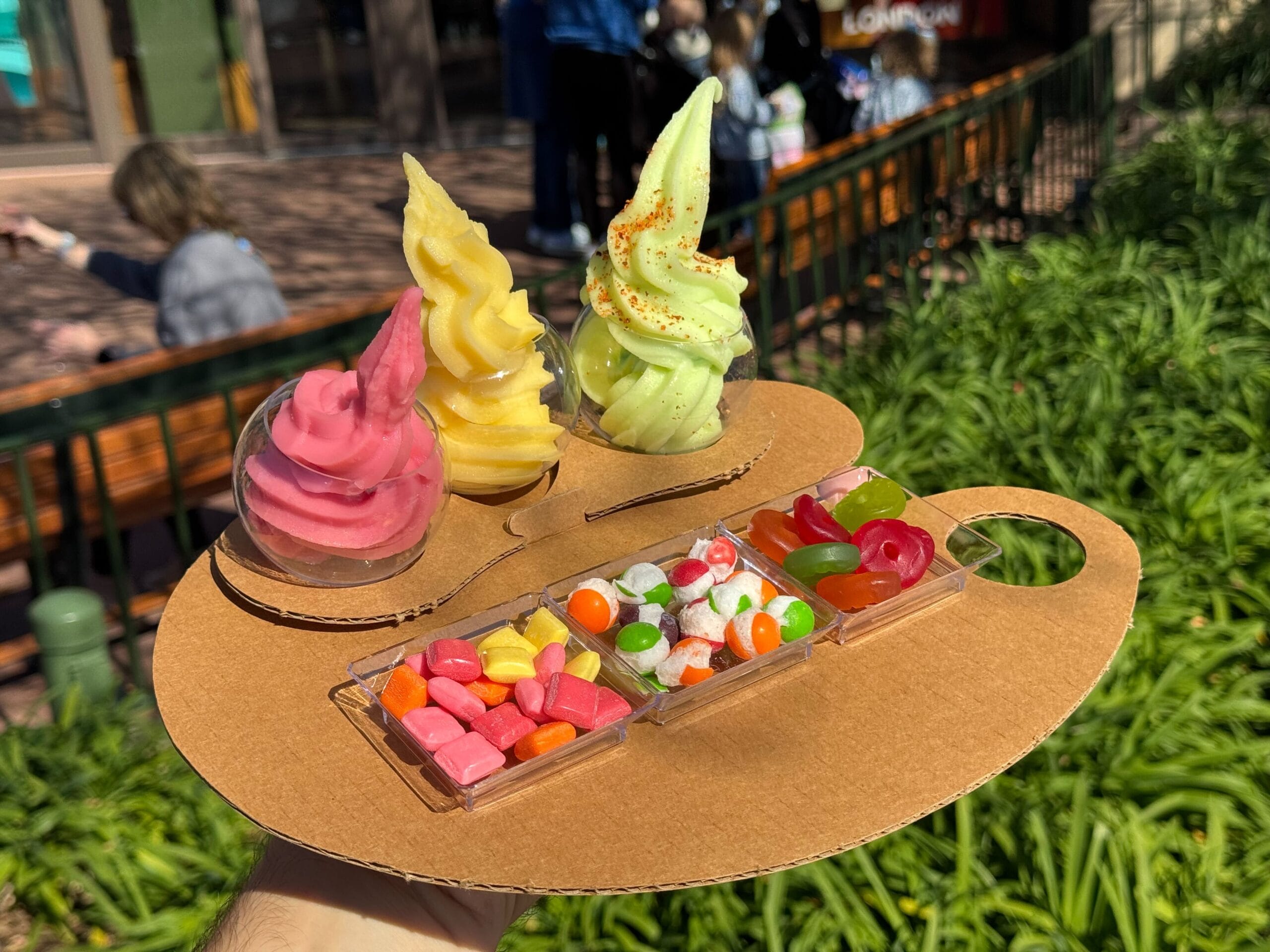 A hand holds a cardboard tray with three swirl ice creams and an assortment of colorful candies in separate compartments, capturing the whimsical spirit of the Festival of the Arts.