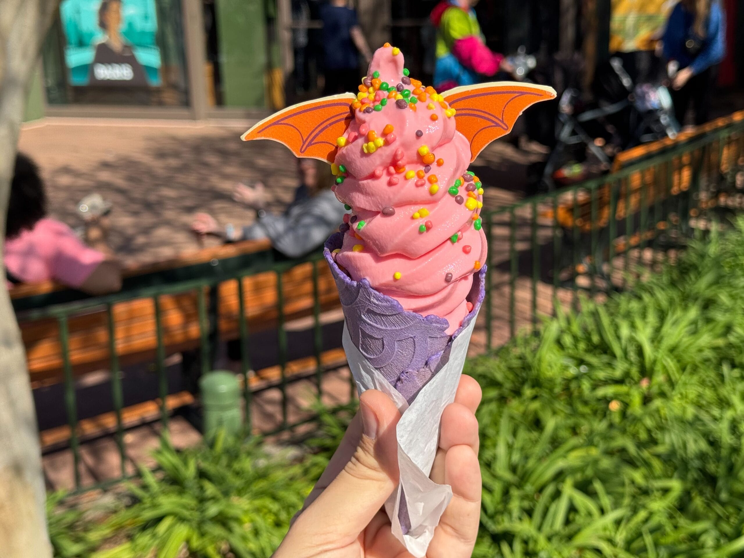 A pink soft-serve ice cream with colorful sprinkles and orange bat wings in a purple cone, held outdoors during the Festival of the Arts, adds a whimsical touch to the park setting.