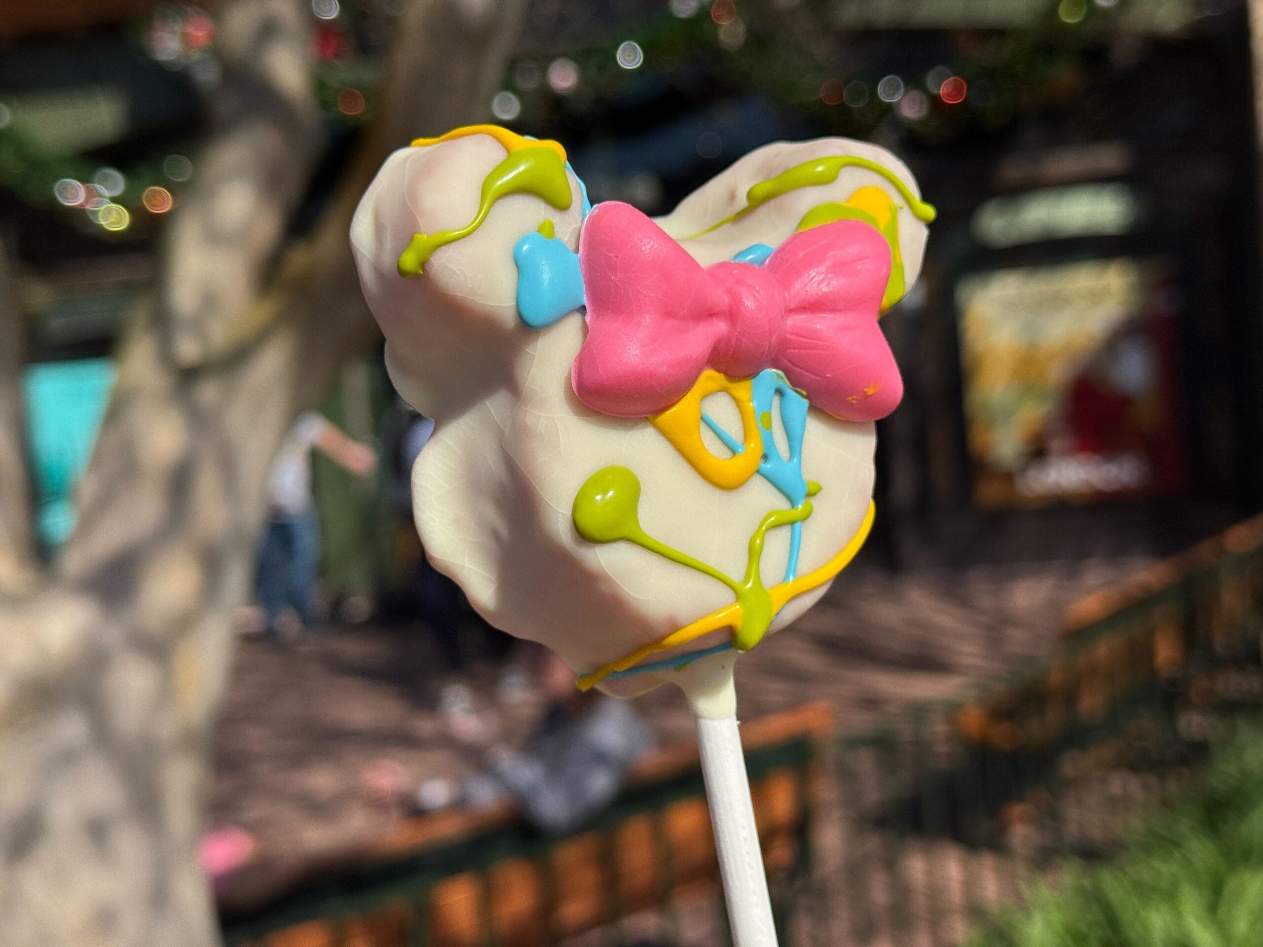 A candy designed like an animal figure with a pink bow, decorated in white, blue, and yellow colors on a stick, held up by a person attending the Festival of the Arts outdoors.