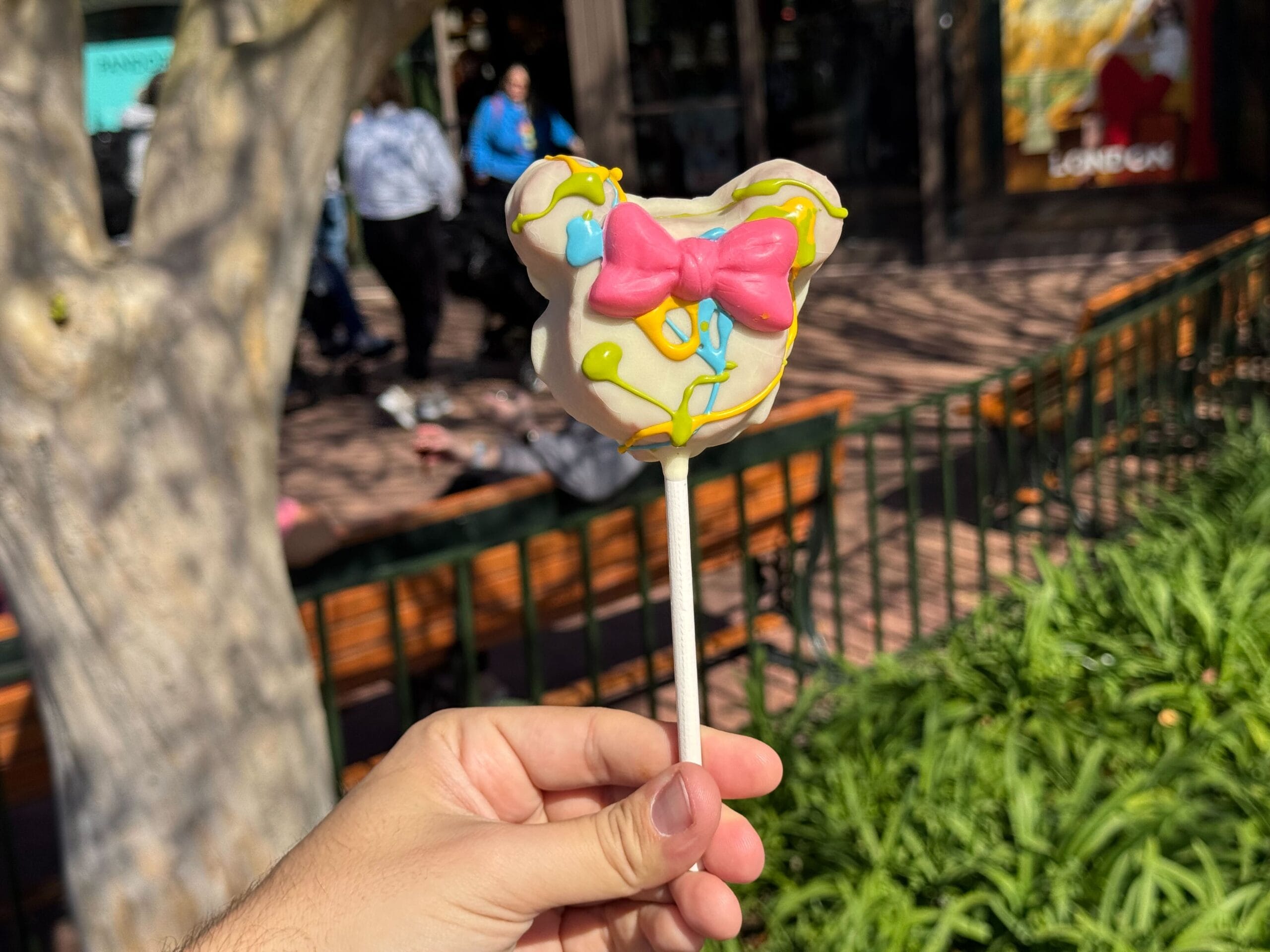 A hand presents a Mickey-shaped cake pop adorned with white icing, a pink bow, and colorful drizzle, capturing the festive spirit of the Festival of the Arts. In the background, a tree and people add to the lively atmosphere.