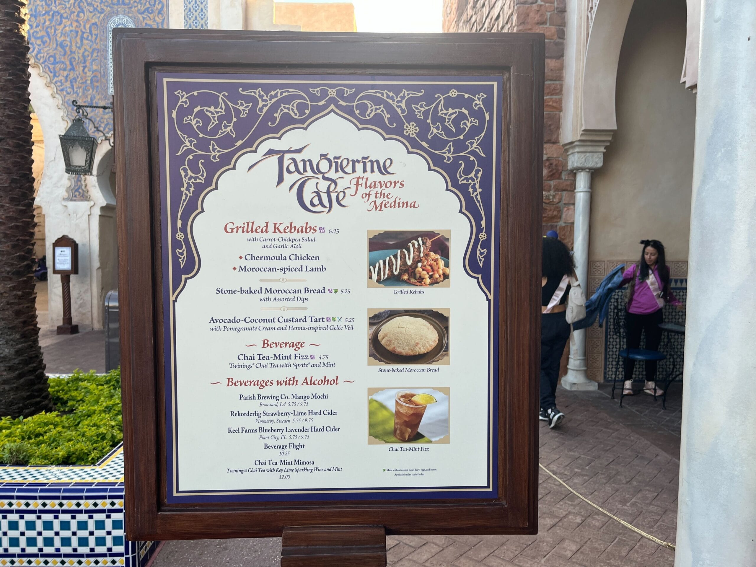 A festival of flavors awaits on the menu board outside our café, showcasing grilled kebabs, avocado couscous tartar, refreshing beverages, and delectable desserts. Framed with a decorative blue and gold border reminiscent of a Festival of the Arts masterpiece.