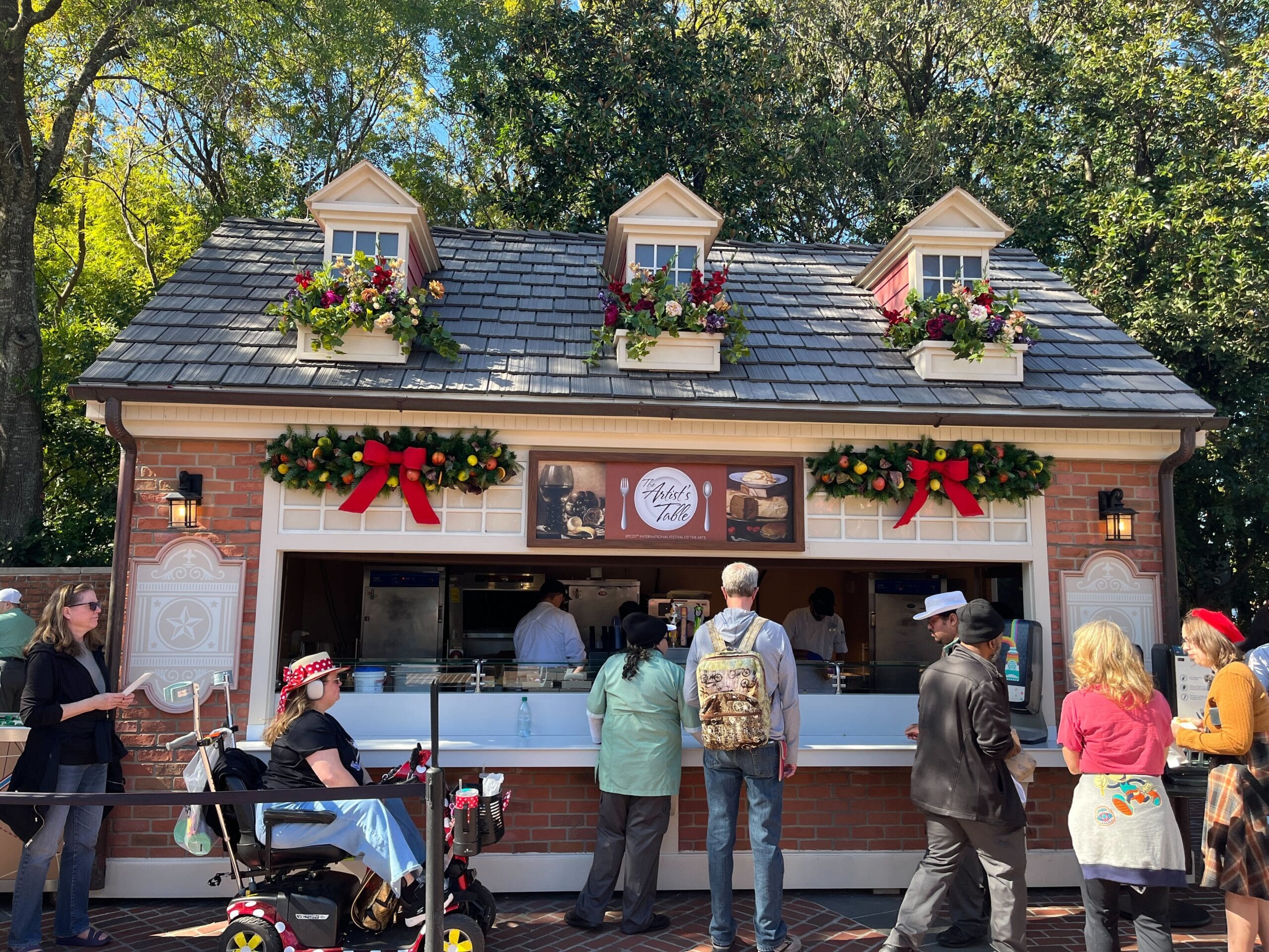 People stand in line at an outdoor food kiosk, decorated with festive garlands and bows, during the Festival of the Arts. The charming brick facade and small displays above the service windows create a lively atmosphere.