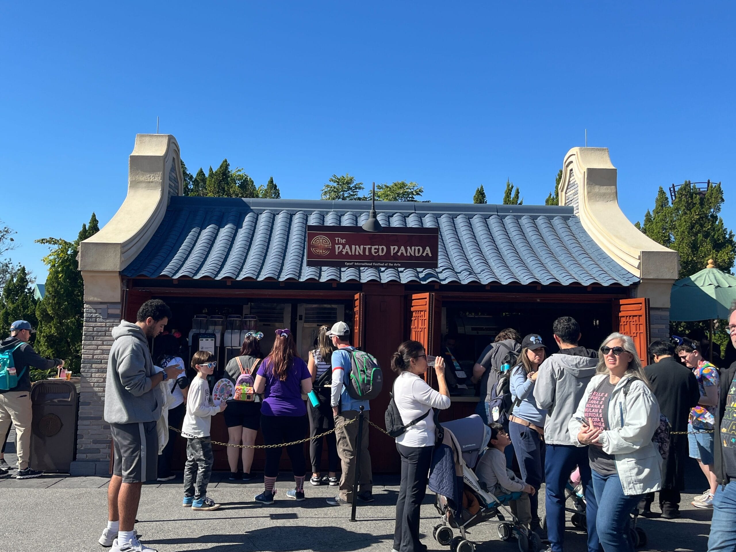People gather around "The Painted Panda" food booth, showcasing Asian-themed architecture, under the sunny sky during the Festival of the Arts. Some wait in line while others interact nearby, soaking up the vibrant atmosphere of culture and creativity.
