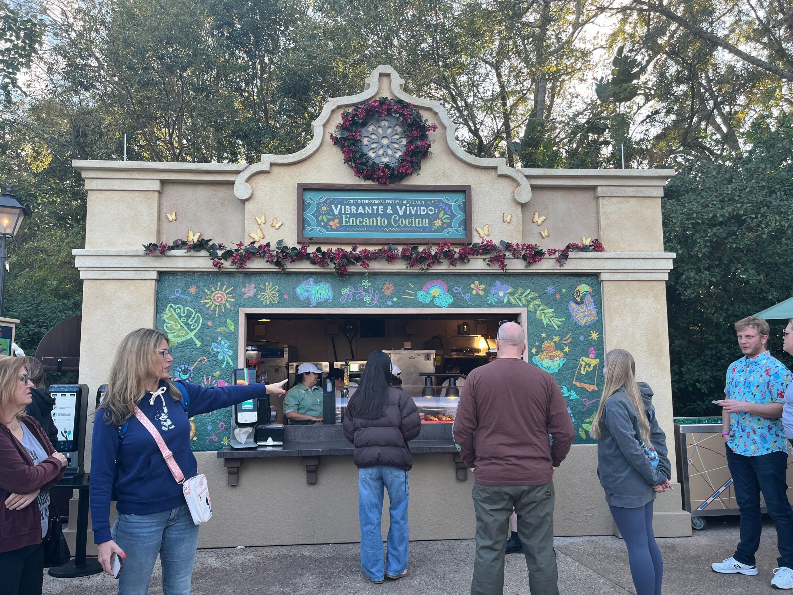 People eagerly wait in line at the Vibrante & Vívido: Encanto Cocina food booth, adorned with floral accents, a delightful highlight of the Festival of the Arts.